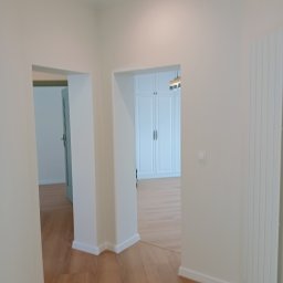 ADEXBUD Usługi Remontowo-Budowlane - Jasne wnętrze po remoncie: drewniana podłoga, białe ściany i zabudowa szaf w tle, widok przez dwa przejścia. Minimalistyczny design, świeżo po wykończeniu.
