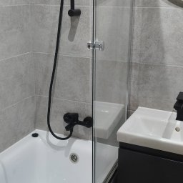 ADEXBUD Usługi Remontowo-Budowlane - Nowoczesna łazienka: biała wanna z czarną armaturą, szklany parawan, umywalka z szafką w odcieniach szarości i okrągłe lustro. Minimalistyczny design i stonowana kolorystyka.