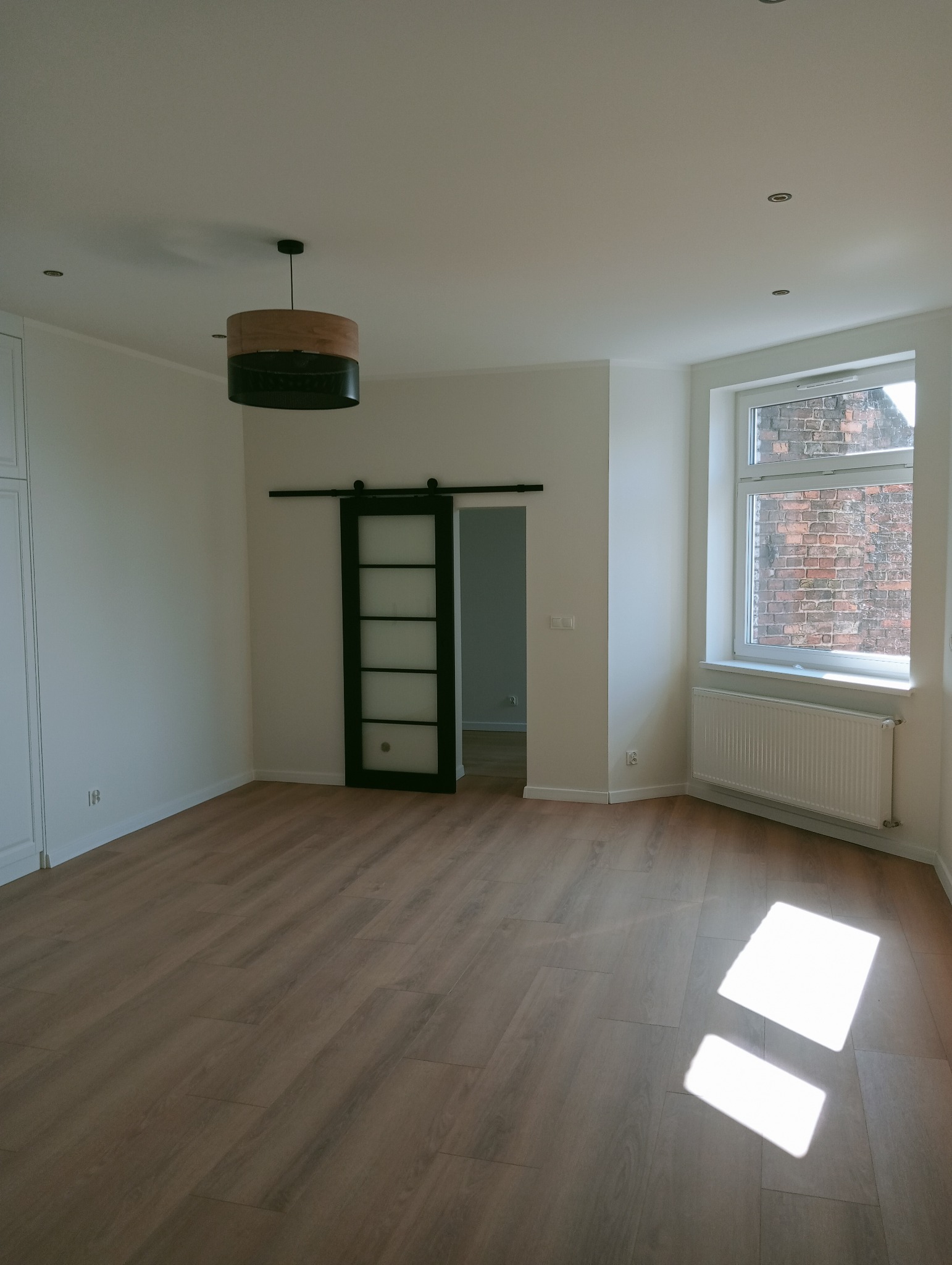 Jasne wnętrze po remoncie: panele podłogowe, białe ściany i okno z widokiem na ceglany budynek. Przesuwne drzwi z czarną ramą i drewniany żyrandol dodają nowoczesnego charakteru.