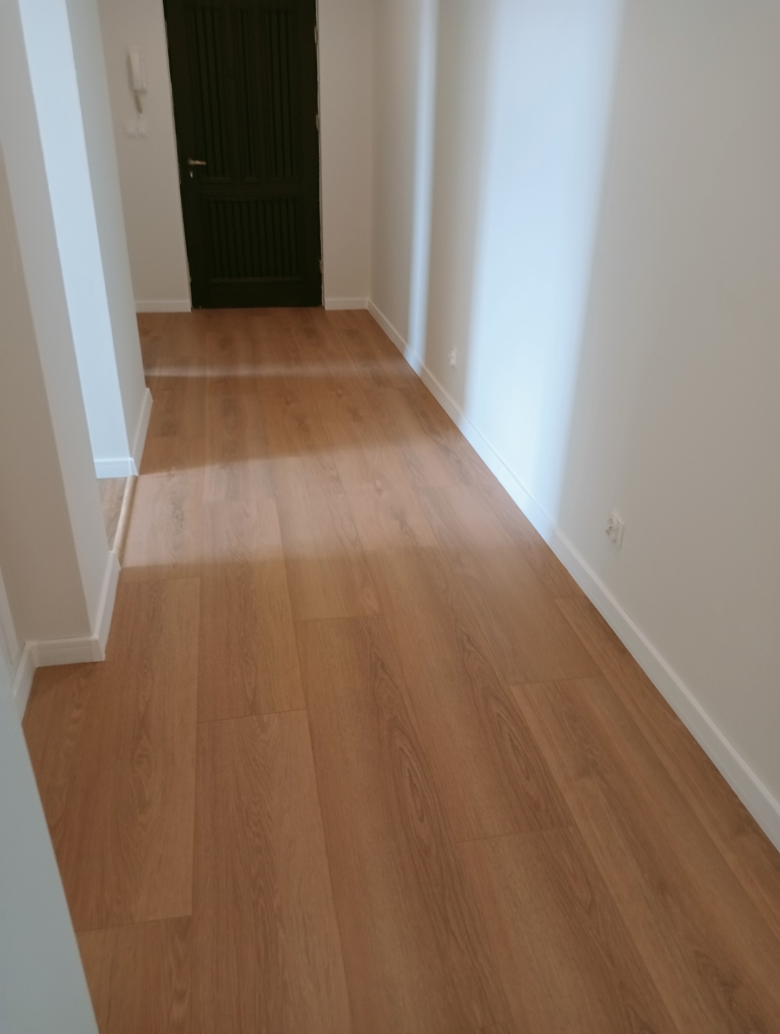 Jasne panele podłogowe w korytarzu z ciemnymi drzwiami. Nowoczesne wykończenie wnętrza z widocznymi listwami przypodłogowymi i gładkimi, białymi ścianami.