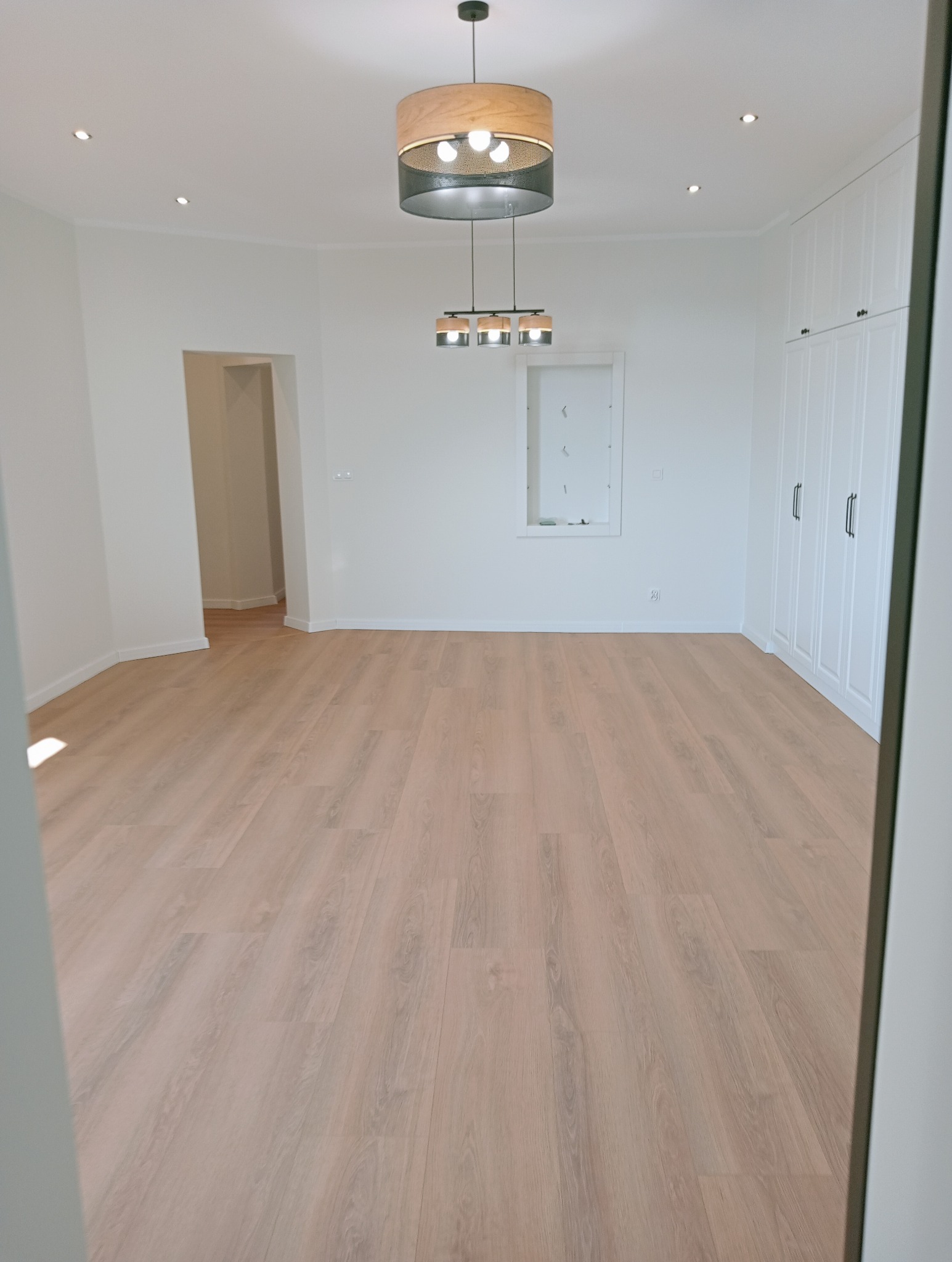 Jasne panele podłogowe w przestronnym pokoju z białymi ścianami, szafą wnękową i nowoczesnym oświetleniem sufitowym. Minimalistyczny design.