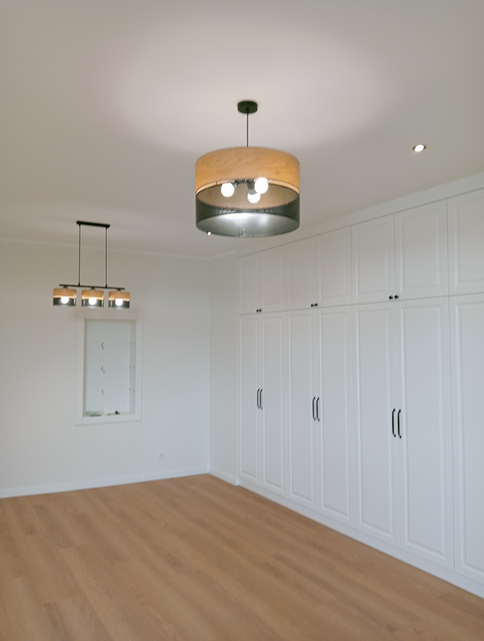 Jasne wnętrze pokoju z panelami podłogowymi i białą zabudową szaf. Dwie lampy sufitowe oświetlają przestrzeń. Minimalistyczny design.