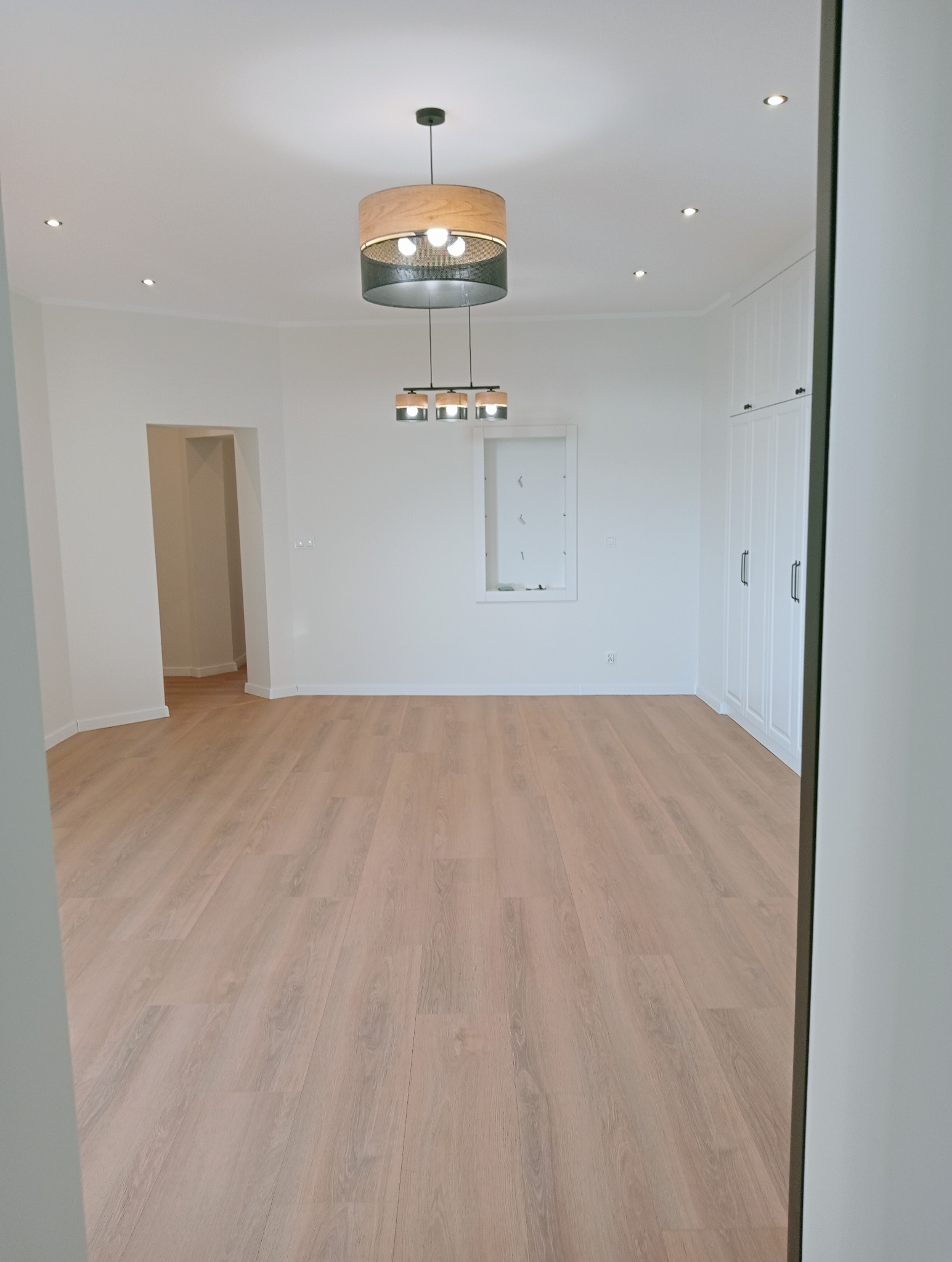 Jasne panele podłogowe w pustym, odświeżonym pokoju z białymi ścianami, wbudowaną szafą i nowoczesnym oświetleniem sufitowym. Widok zza framugi.