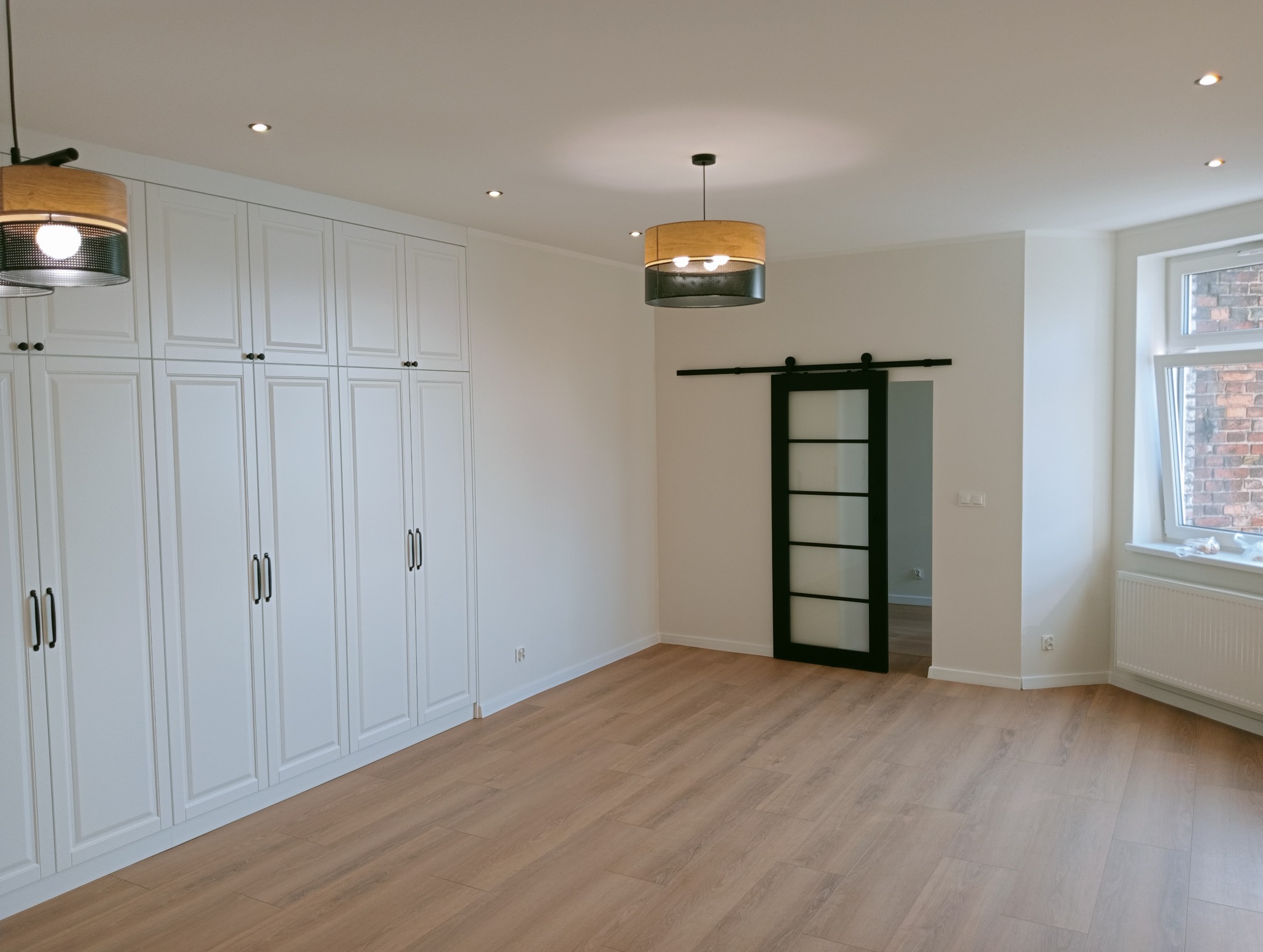 Jasne wnętrze po remoncie: drewniana podłoga, biała zabudowa szaf, przesuwne drzwi z czarną ramą i okno z widokiem na ceglany budynek.