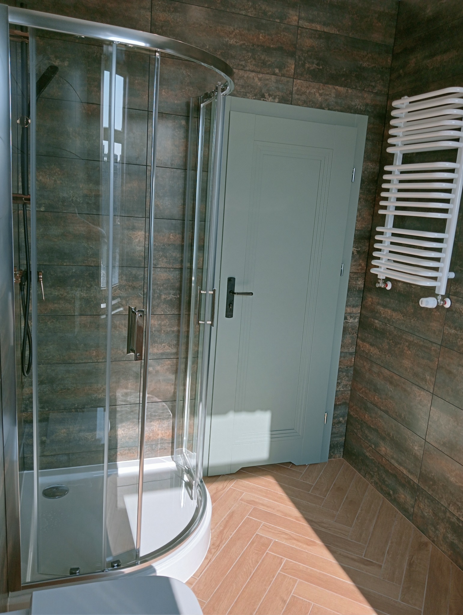 Nowoczesna kabina prysznicowa z chromowanymi detalami, szklanymi drzwiami i jasną, drewnopodobną podłogą ułożoną w jodełkę. W tle ciemne płytki i grzejnik.