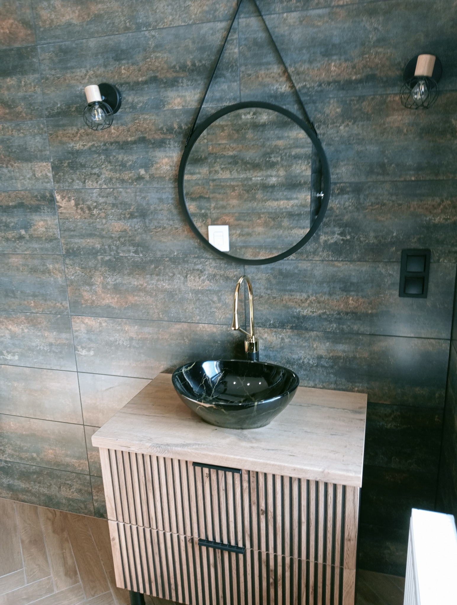 Stylowa łazienka z umywalką z czarnego marmuru, okrągłym lustrem na tle płytek imitujących rdzę i drewnianą szafką z frezowanymi frontami.
