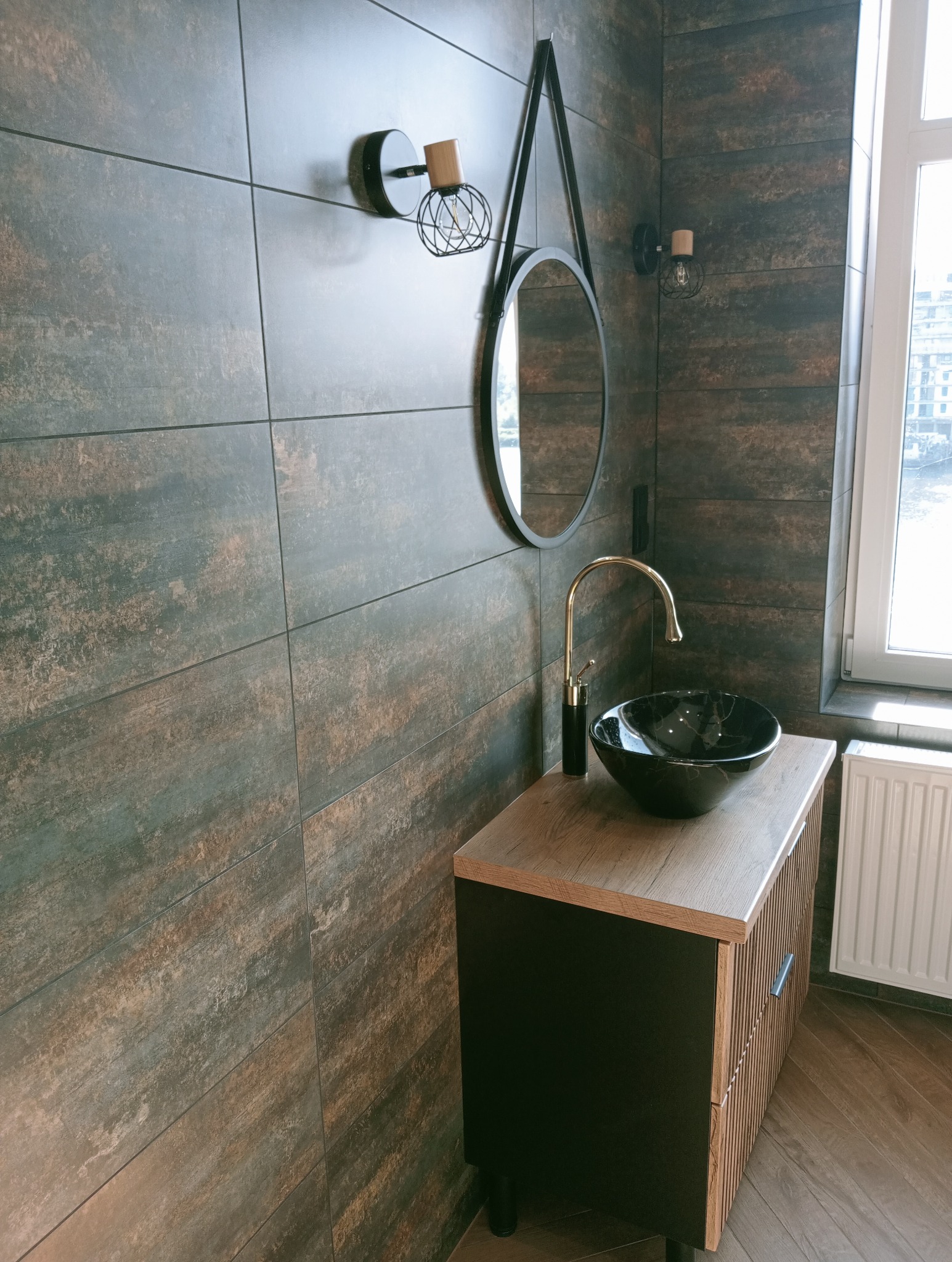 Nowoczesna łazienka z umywalką z czarnego marmuru i złotą armaturą na tle płytek imitujących rdzę. Owalne lustro i designerskie lampy dodają elegancji.