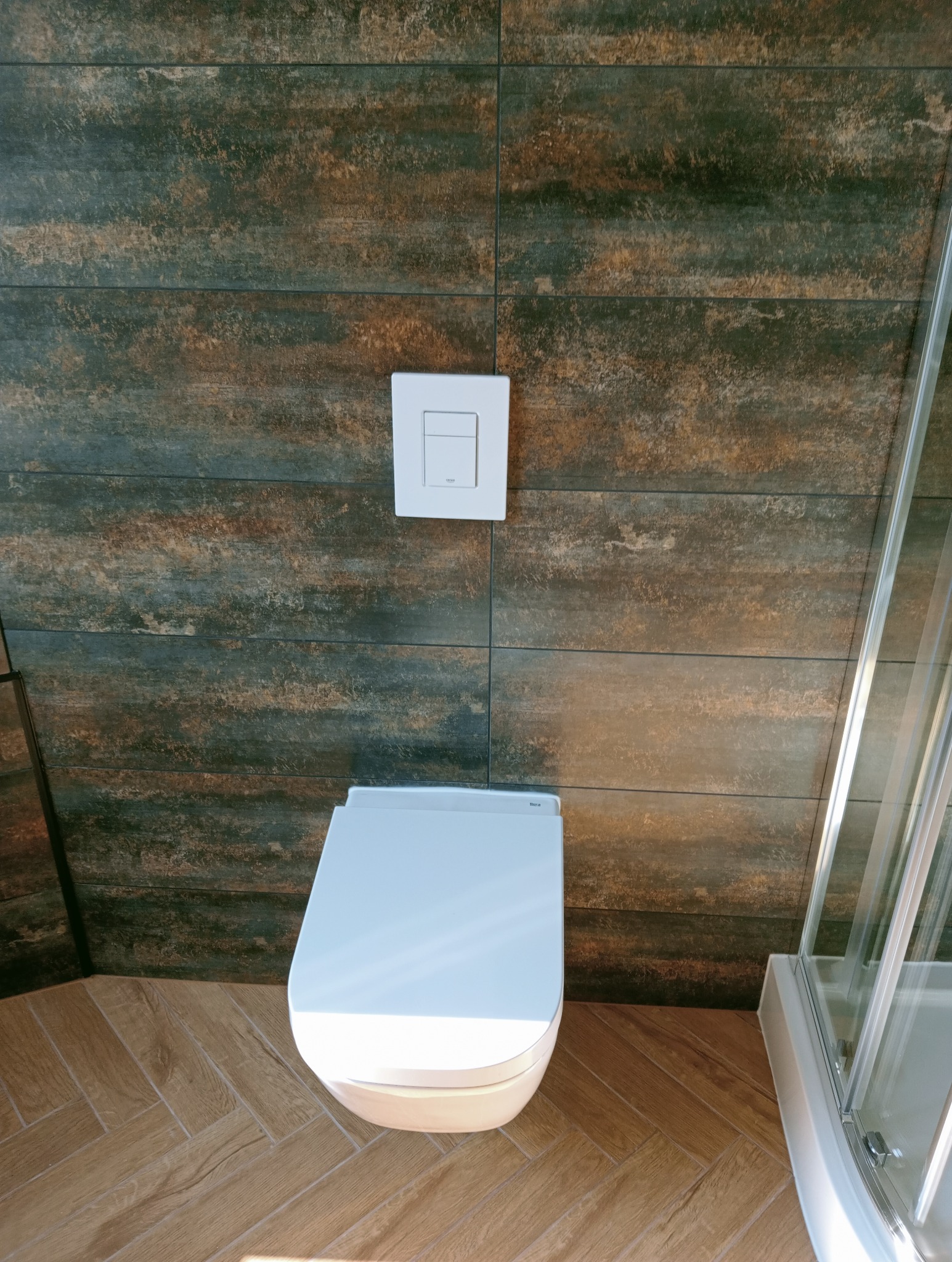 Nowoczesna toaleta podwieszana z białym przyciskiem spłukującym na tle płytek imitujących drewno i fragmentu kabiny prysznicowej. Parkiet ułożony w jodełkę.