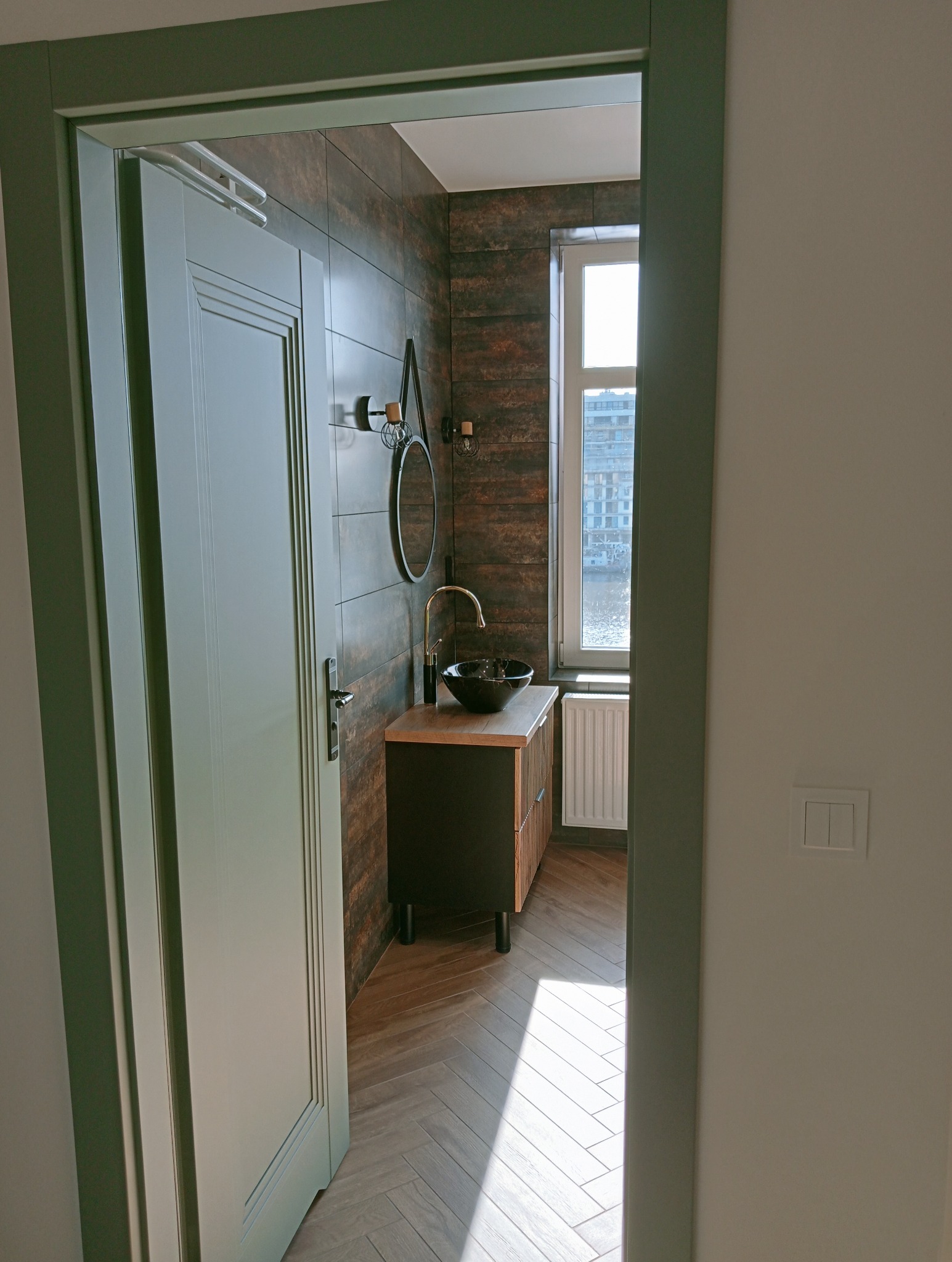 Nowoczesna łazienka z brązowymi płytkami, czarną umywalką i drewnianym blatem, widziana zza otwartych drzwi w kolorze stonowanej zieleni. Parkiet ułożony w jodełkę.
