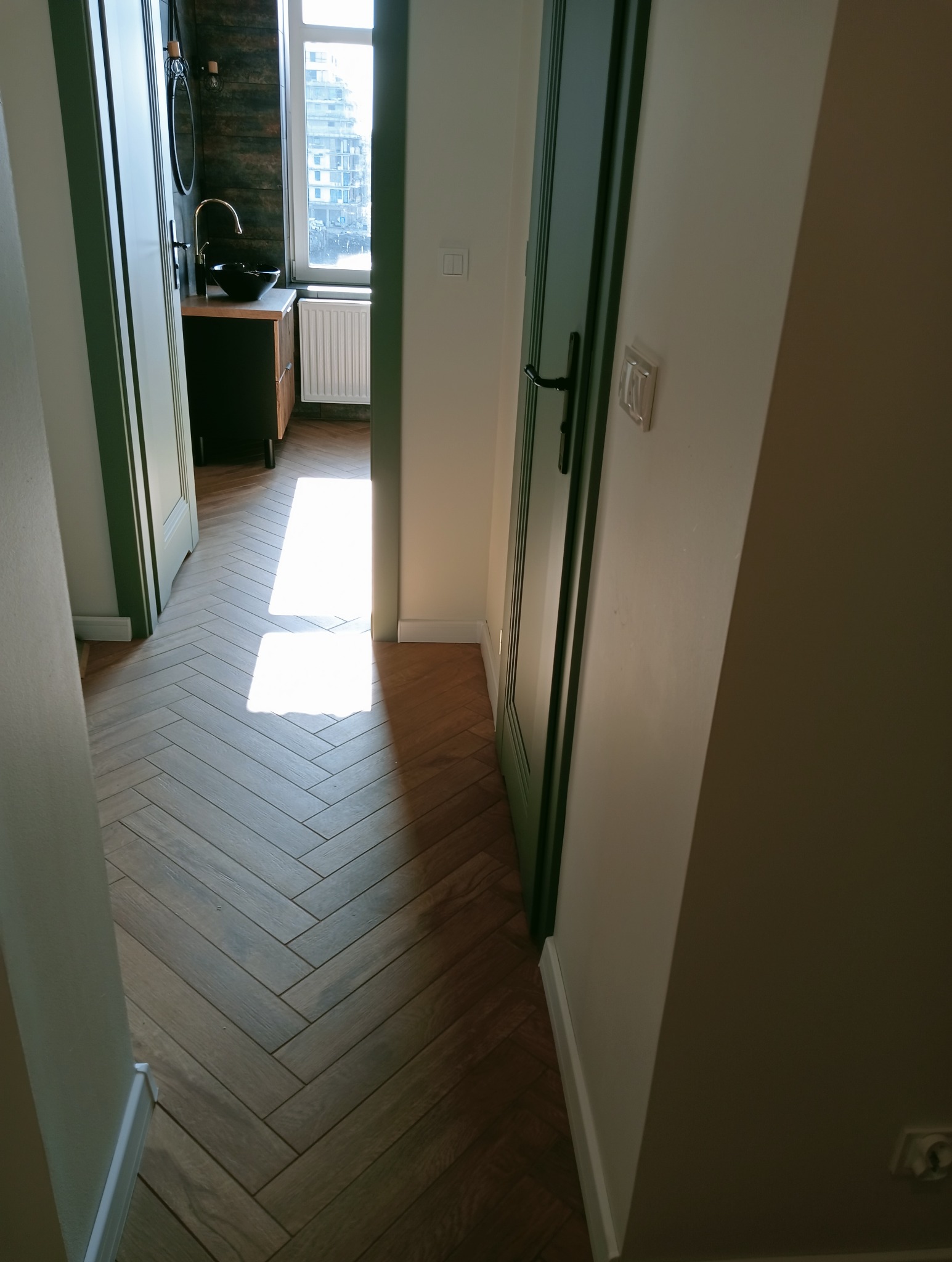 Wnętrze z parkietem w jodełkę, widok na łazienkę z umywalką na szafce i oknem. Drzwi w kolorze butelkowej zieleni. Nowoczesny styl.