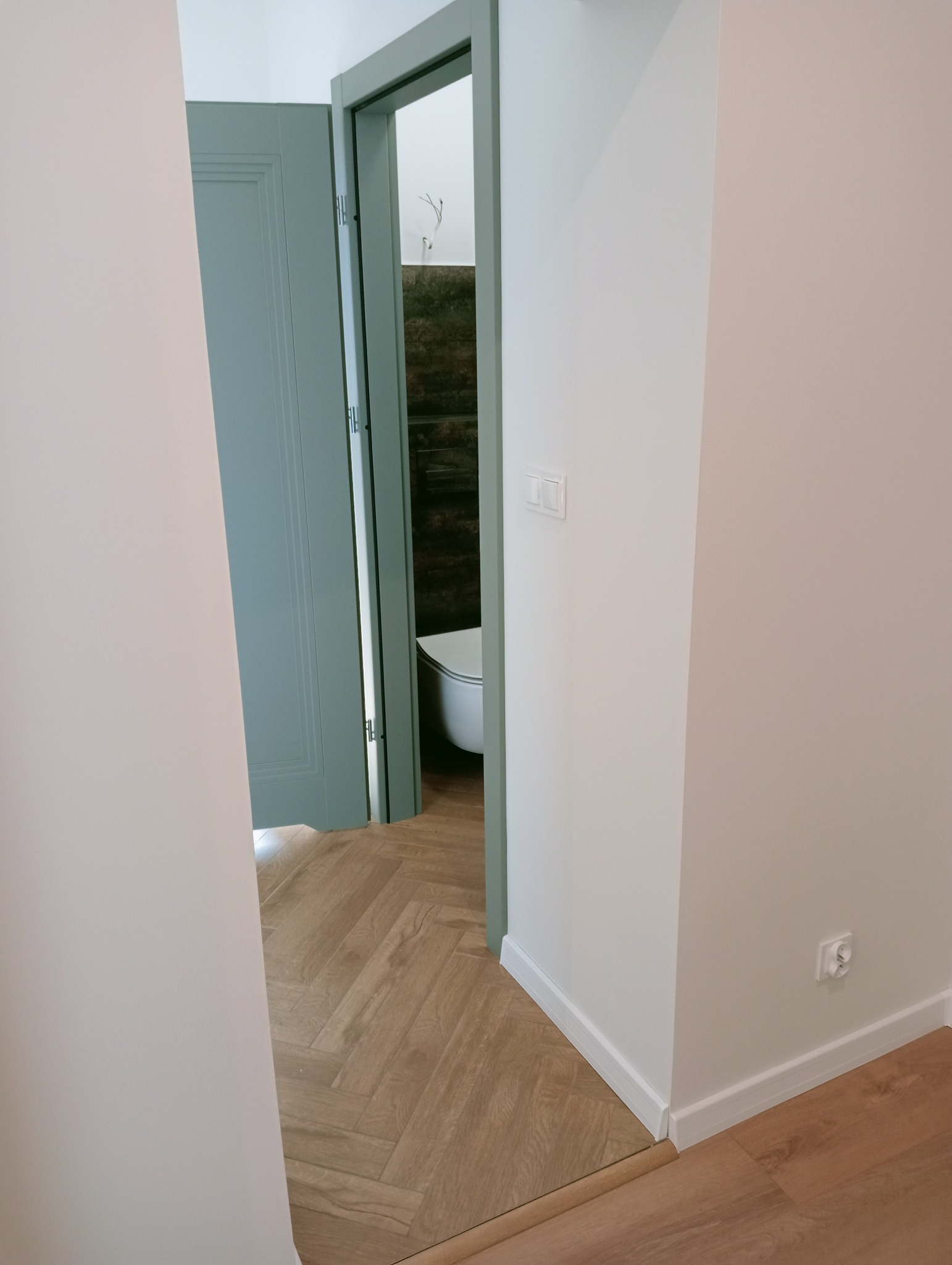 Jasne wnętrze po remoncie: parkiet w jodełkę, białe ściany, niebieskie drzwi do łazienki z brązowymi płytkami. Widoczny fragment toalety.