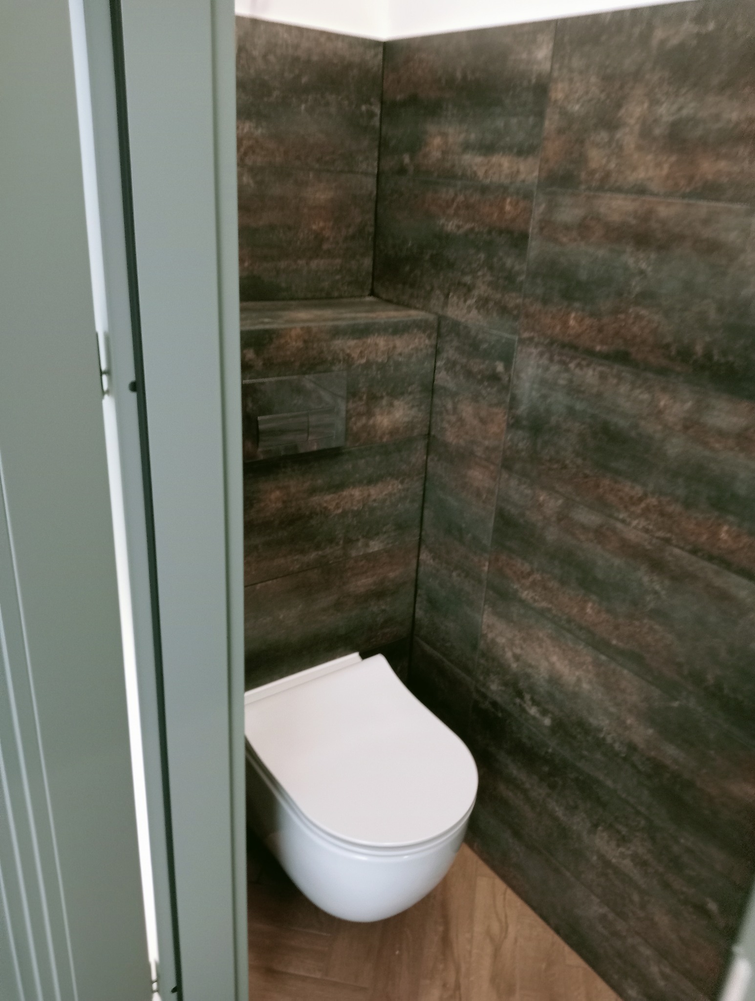 Nowoczesna toaleta podwieszana z brązowymi płytkami imitującymi drewno, widoczna zza uchylonych, szarych drzwi. Ukończony remont łazienki.