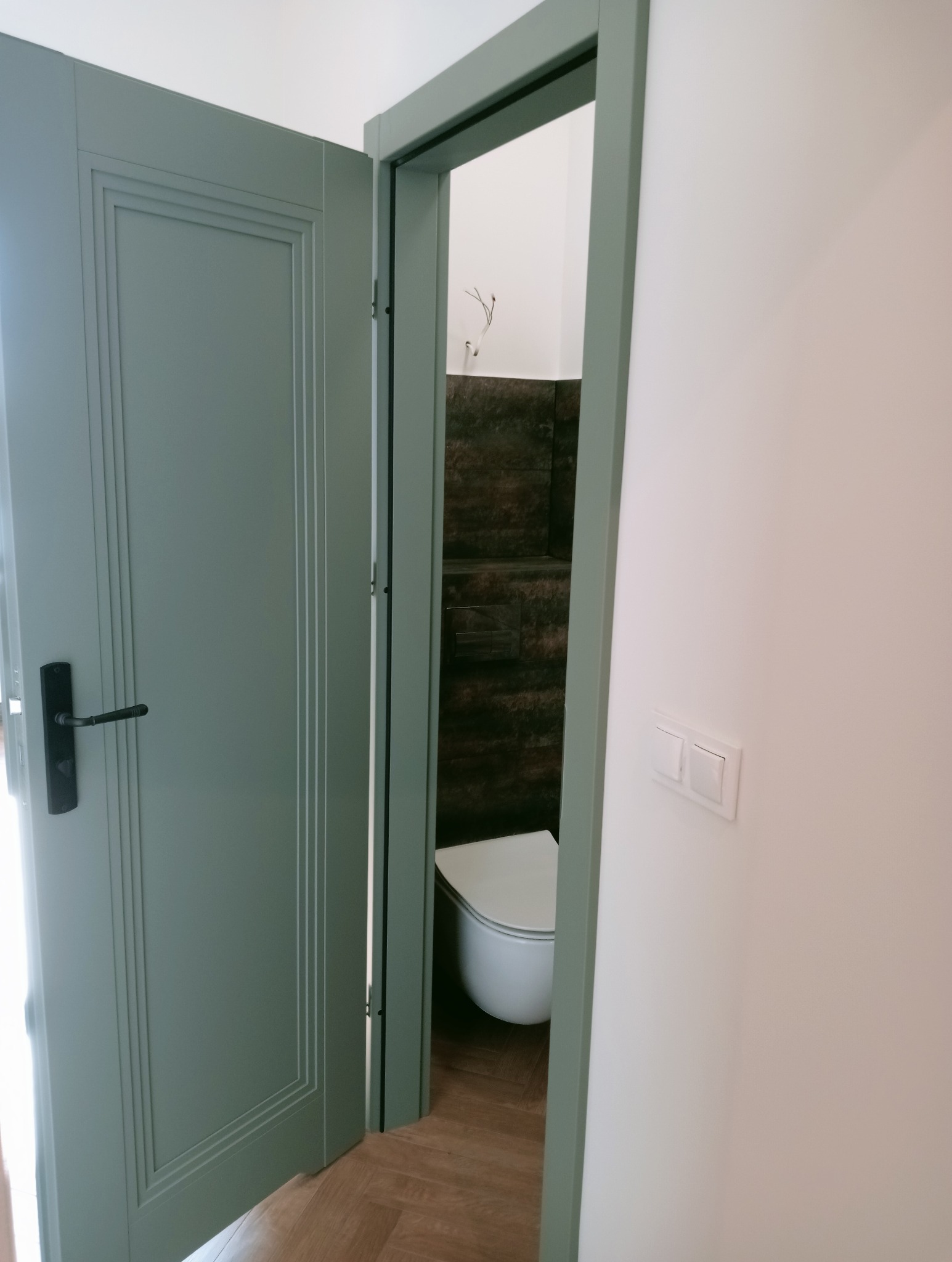 Otwarta, pastelowa, zielona  rama drzwiowa prowadzi do wyremontowanej łazienki z brązowymi płytkami i podłogą w jodełkę. Widoczny biały sedes i włącznik światła.