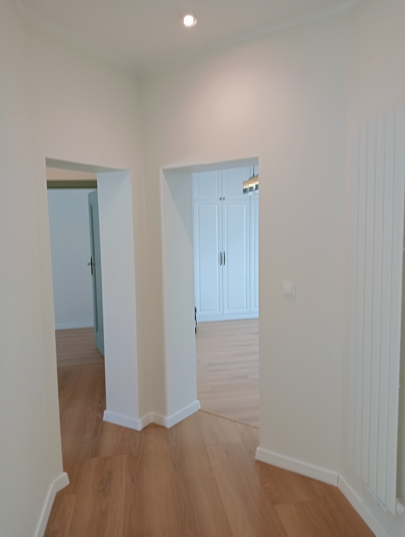 Jasne wnętrze po remoncie: drewniana podłoga, białe ściany i zabudowa szaf w tle, widok przez dwa przejścia. Minimalistyczny design, świeżo po wykończeniu.