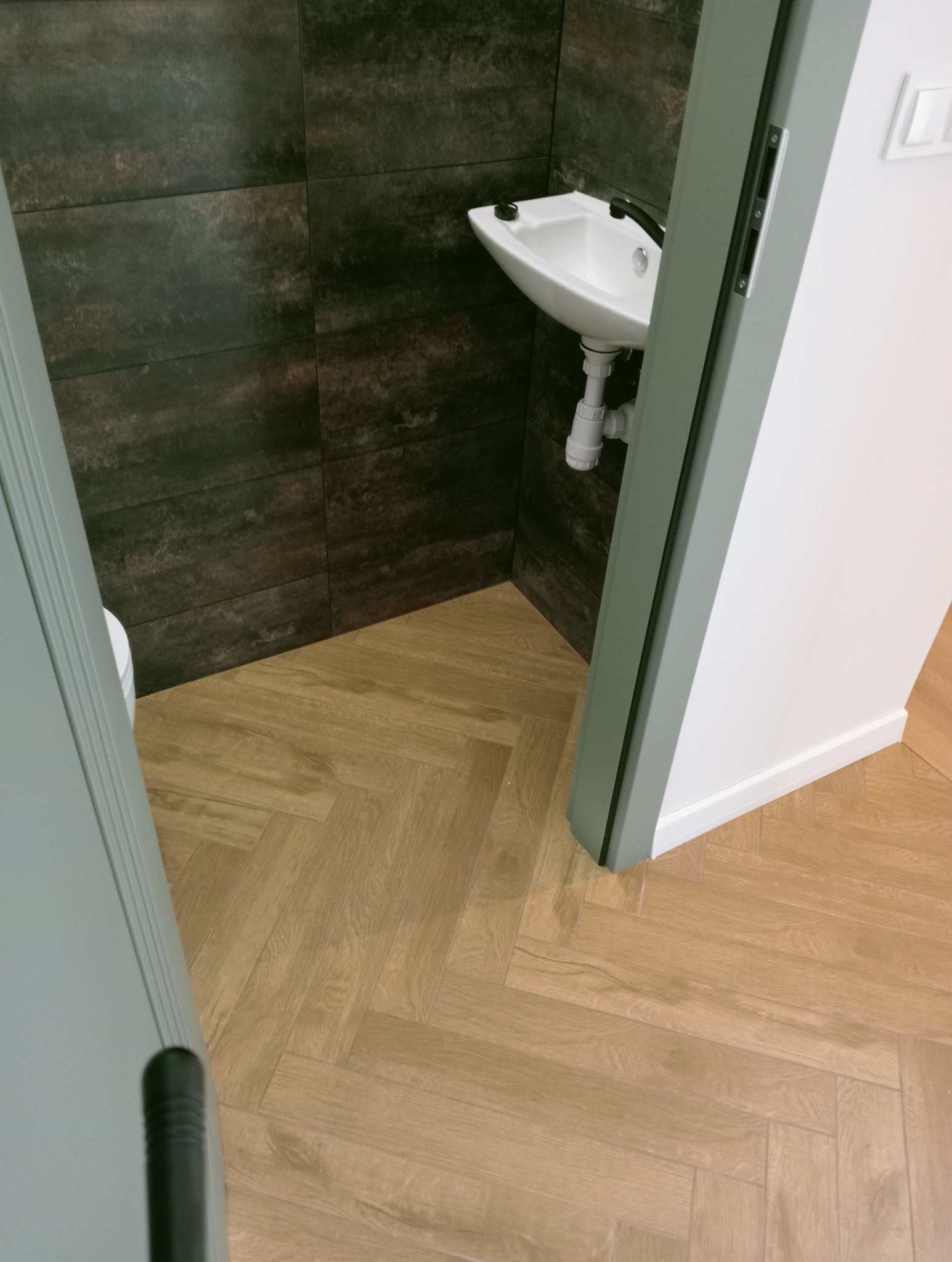 Nowoczesna łazienka po remoncie: ciemne, marmurowe płytki ścienne, biała umywalka i jasna podłoga ułożona w jodełkę. Widok na odnowione wnętrze przez otwarte drzwi.