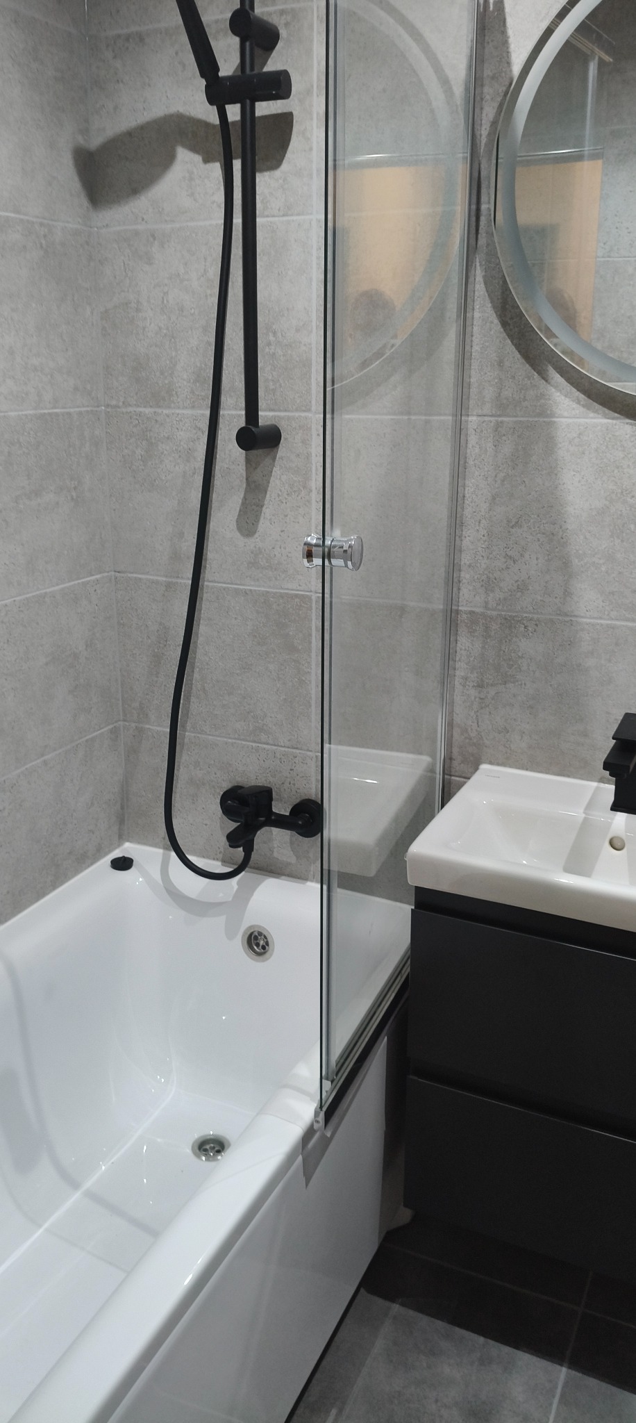 Nowoczesna łazienka: biała wanna z czarną armaturą, szklany parawan, umywalka z szafką w odcieniach szarości i okrągłe lustro. Minimalistyczny design i stonowana kolorystyka.