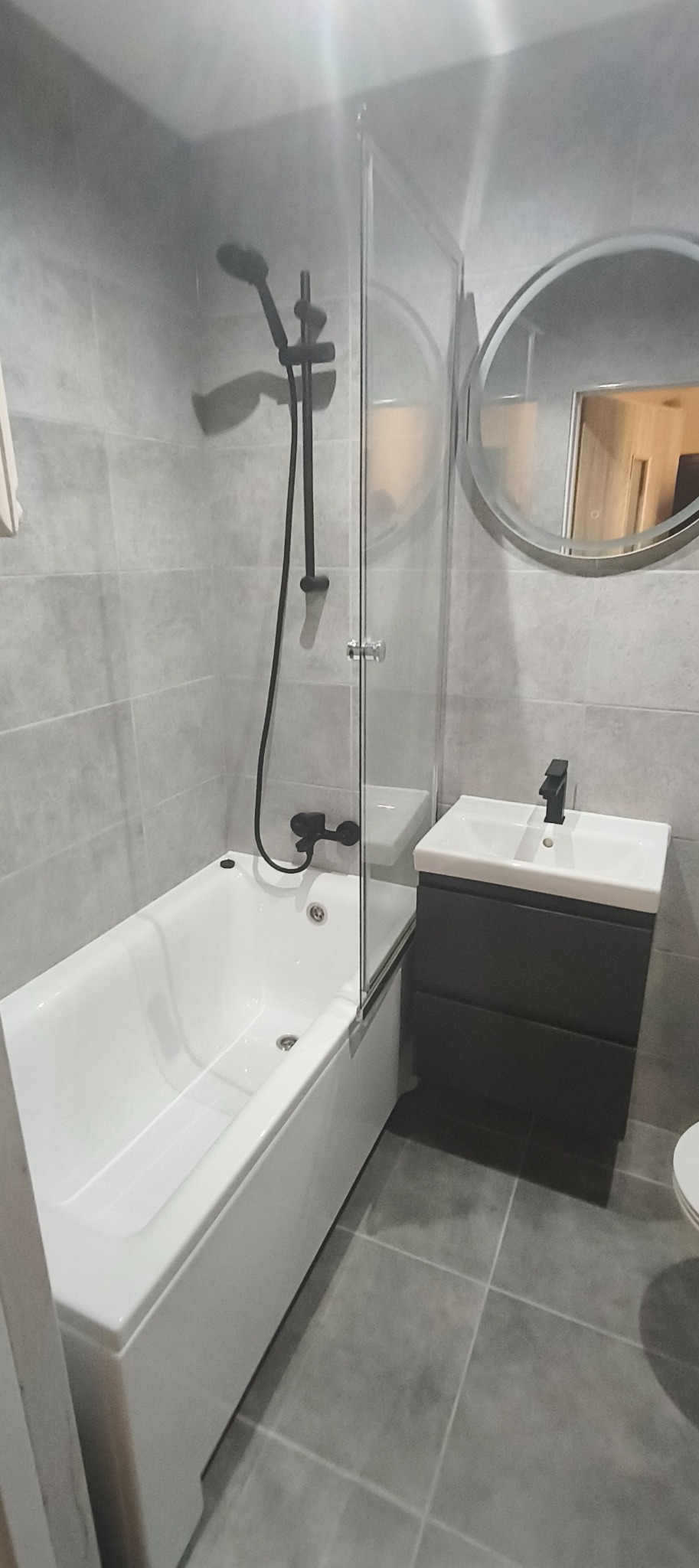 Nowoczesna łazienka w Warnicach: szare płytki, biała wanna z parawanem, czarna armatura i okrągłe lustro nad umywalką. Minimalistyczny design.