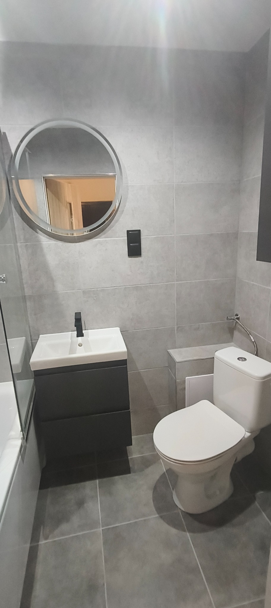 Szara łazienka po remoncie z okrągłym lustrem podświetlanym LED, umywalką z szafką i toaletą. Płytki na ścianach i podłodze w odcieniach szarości.