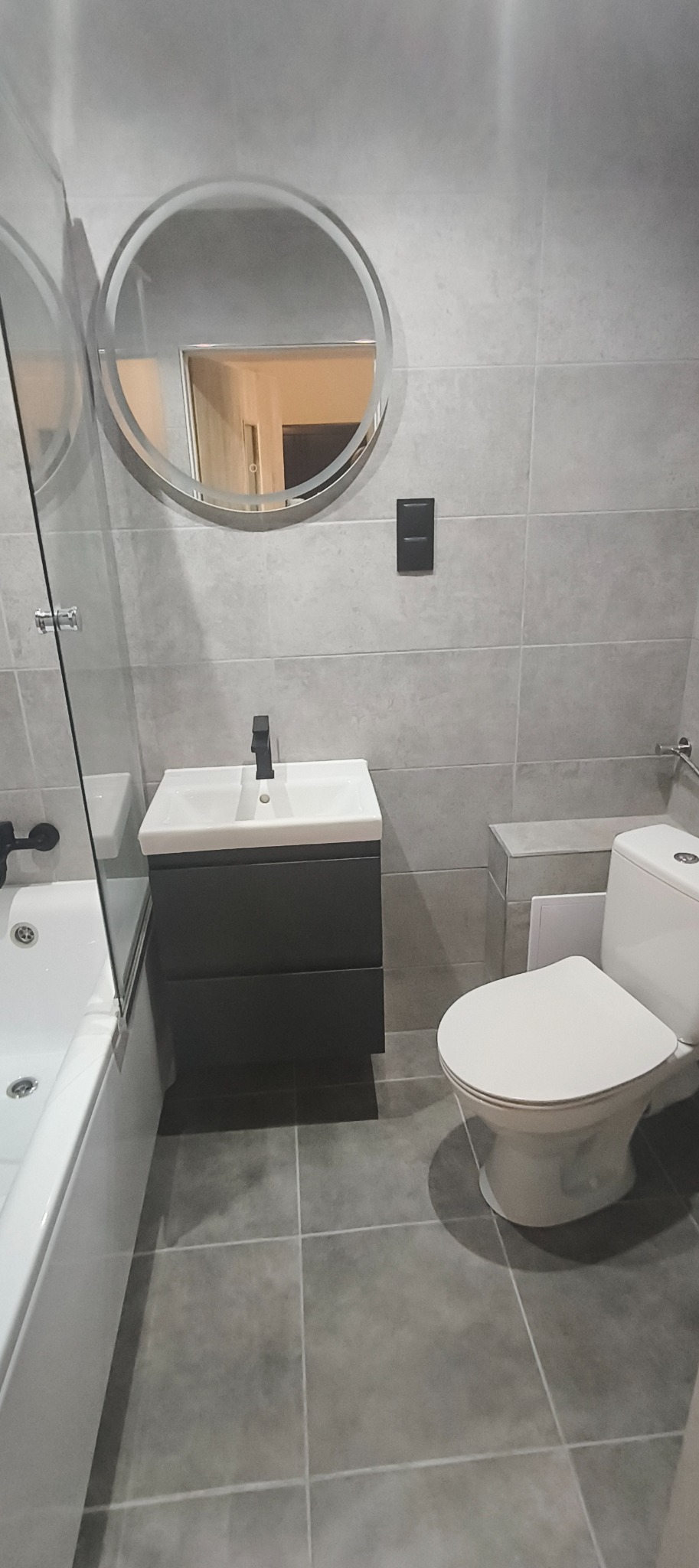 Nowoczesna łazienka w szarościach: okrągłe lustro z podświetleniem, umywalka z szafką w kolorze antracytu, biała toaleta. Minimalistyczny design, płytki w odcieniach szarości.