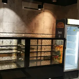 Wnętrze piekarni lub cukierni z witryną chłodniczą i lodówką na napoje, widoczny zamontowany klimatyzator podsufitowy.