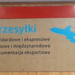 Tablica informacyjna z napisem 'Przesyłki' w kolorze niebieskim, pod spodem tekst 'Standardowe i ekspresowe, Krajowe i międzynarodowe, Dokumentacja eksportowa' oraz niebieska grafika samolotu.