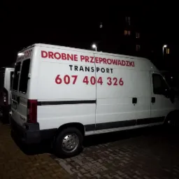 Biały samochód dostawczy z czerwonym napisem 'Drobne Przeprowadzki TRANSPORT 607 404 326' zaparkowany na parkingu w nocy.