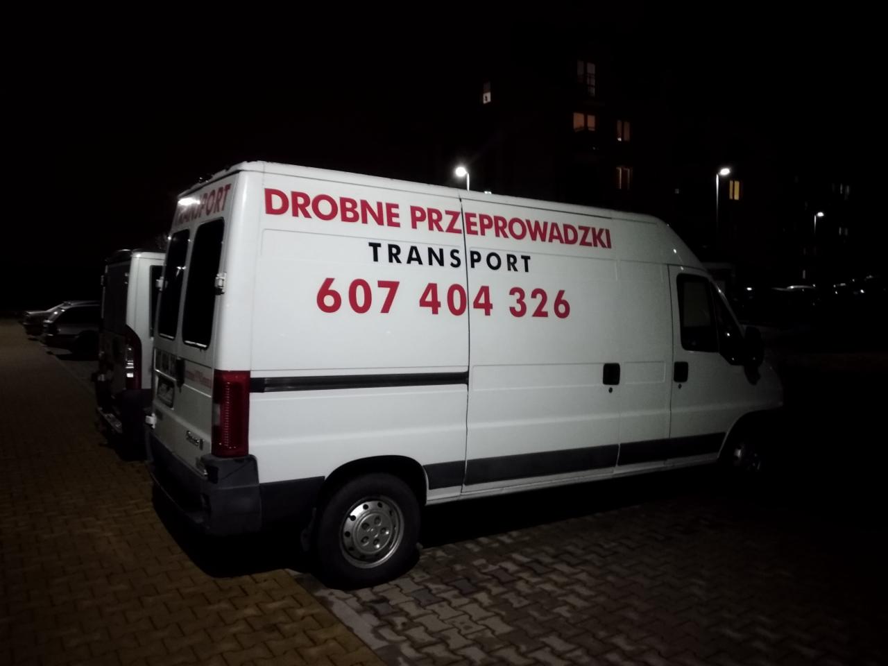 Biały samochód dostawczy z czerwonym napisem 'Drobne Przeprowadzki TRANSPORT 607 404 326' zaparkowany na parkingu w nocy.
