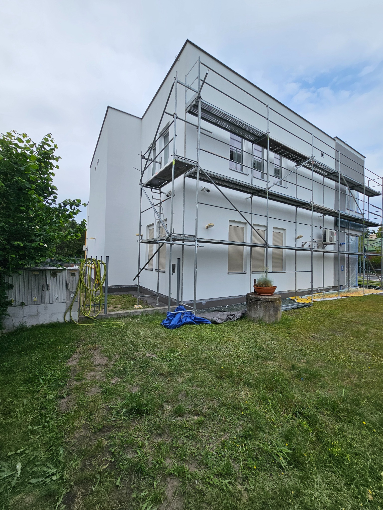 Biały, dwukondygnacyjny dom w trakcie prac elewacyjnych, otoczony rusztowaniem. Widoczne elementy ogrodu i trawnika. W tle pochmurne niebo.