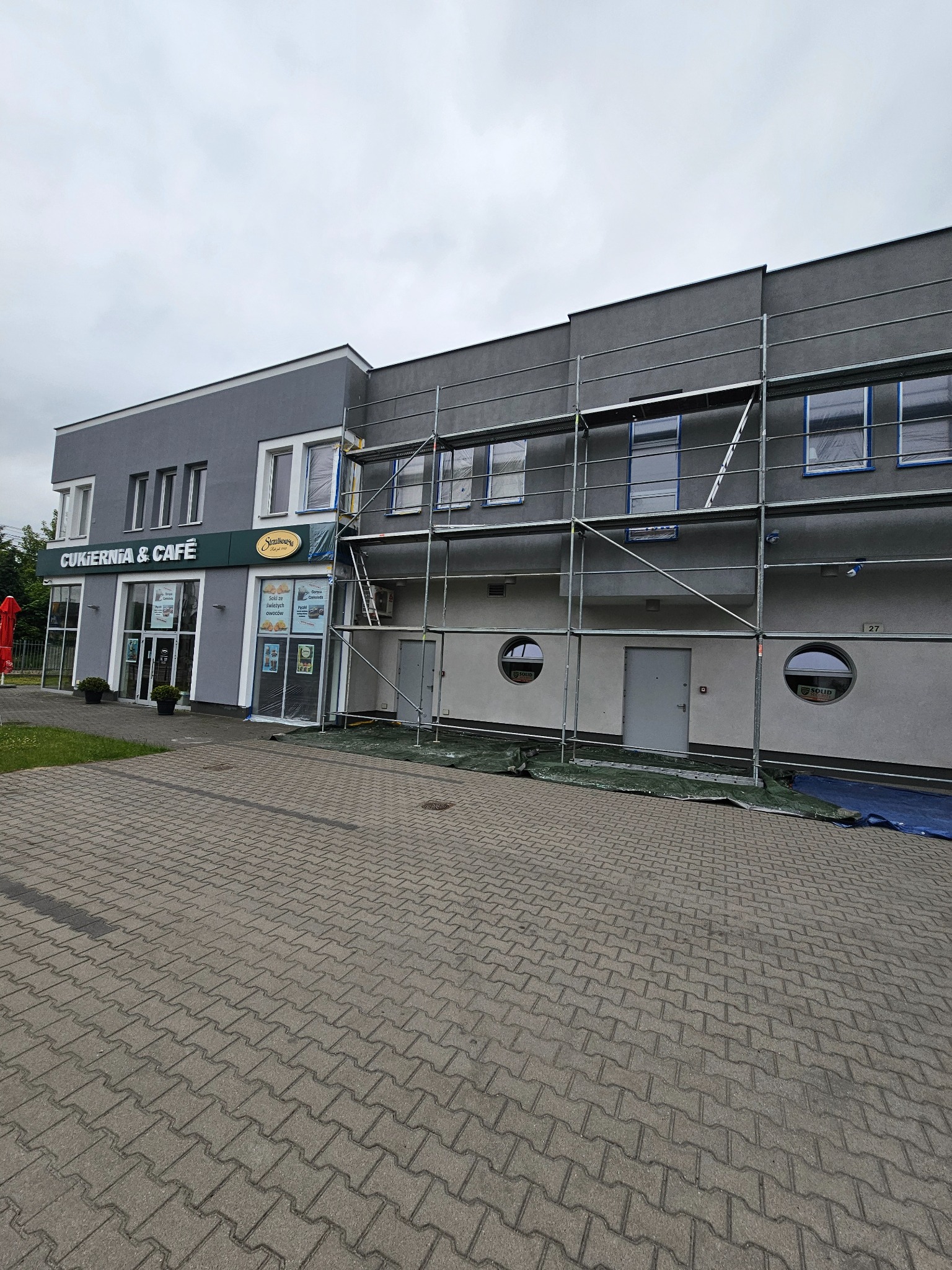 Budynek Cukierni & Cafe w trakcie malowania elewacji, z widocznymi rusztowaniami i zabezpieczonymi oknami. Szara kolorystyka budynku.
