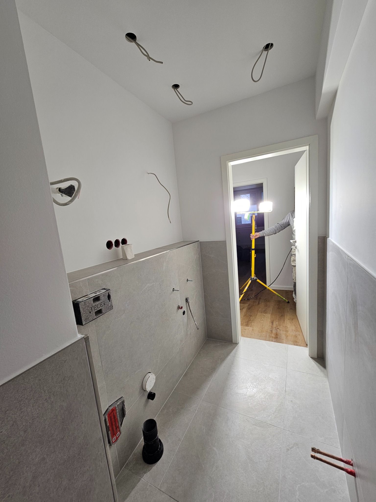 Wnętrze łazienki w trakcie wykończenia: szare płytki na ścianach i podłodze, widoczne instalacje, oświetlenie budowlane. Prace glazurnicze w toku.