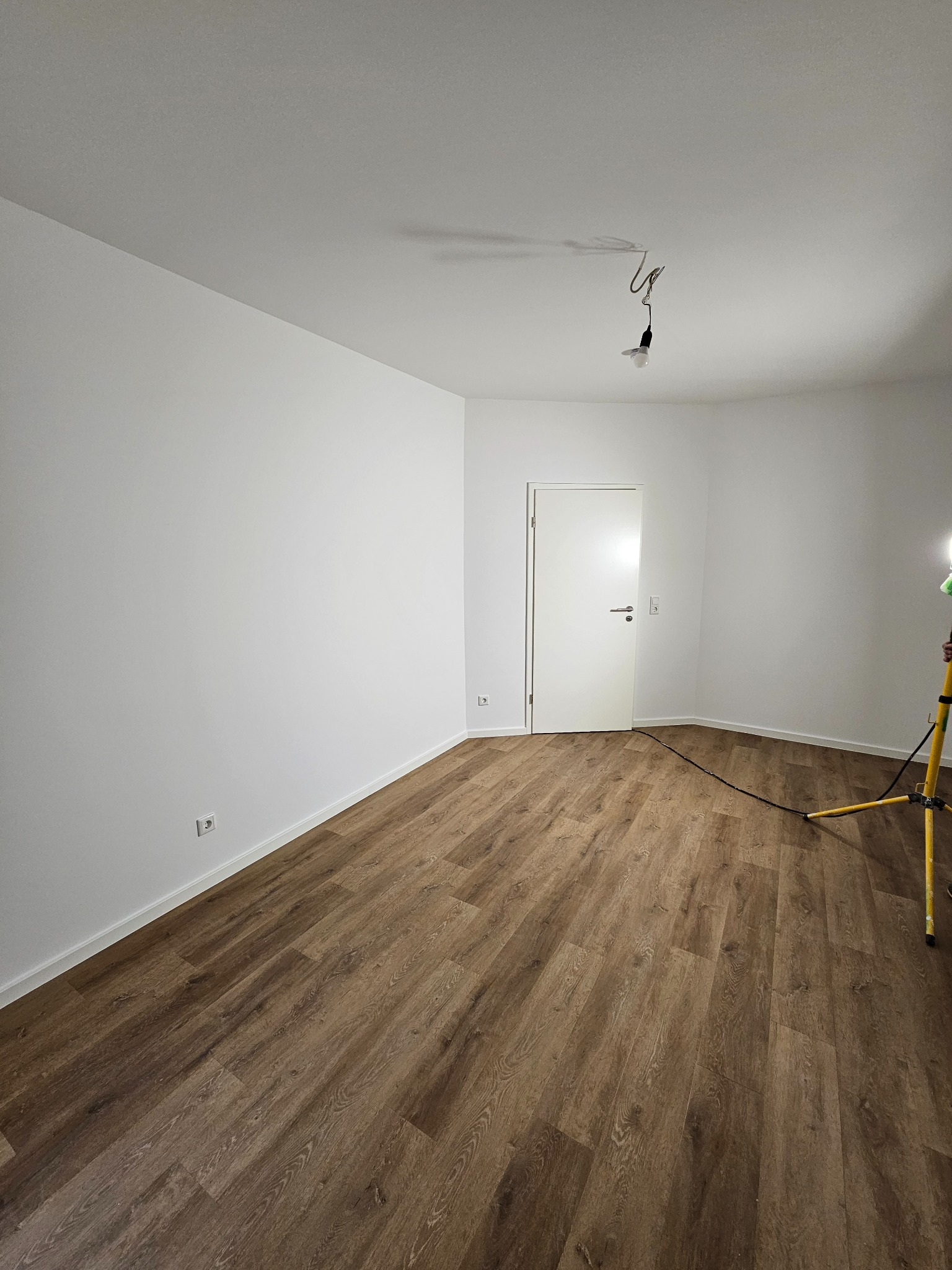 Pusty pokój po remoncie: świeżo pomalowane białe ściany, brązowa podłoga imitująca drewno, białe drzwi. Widoczna lampa robocza na trójnogu.