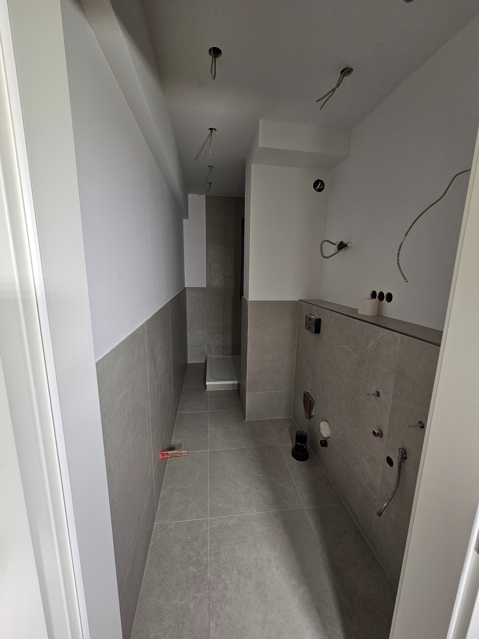 Szara łazienka w trakcie wykańczania: płytki na ścianach i podłodze, widoczne instalacje, surowy stan przed montażem armatury. Minimalistyczny design, neutralna kolorystyka.