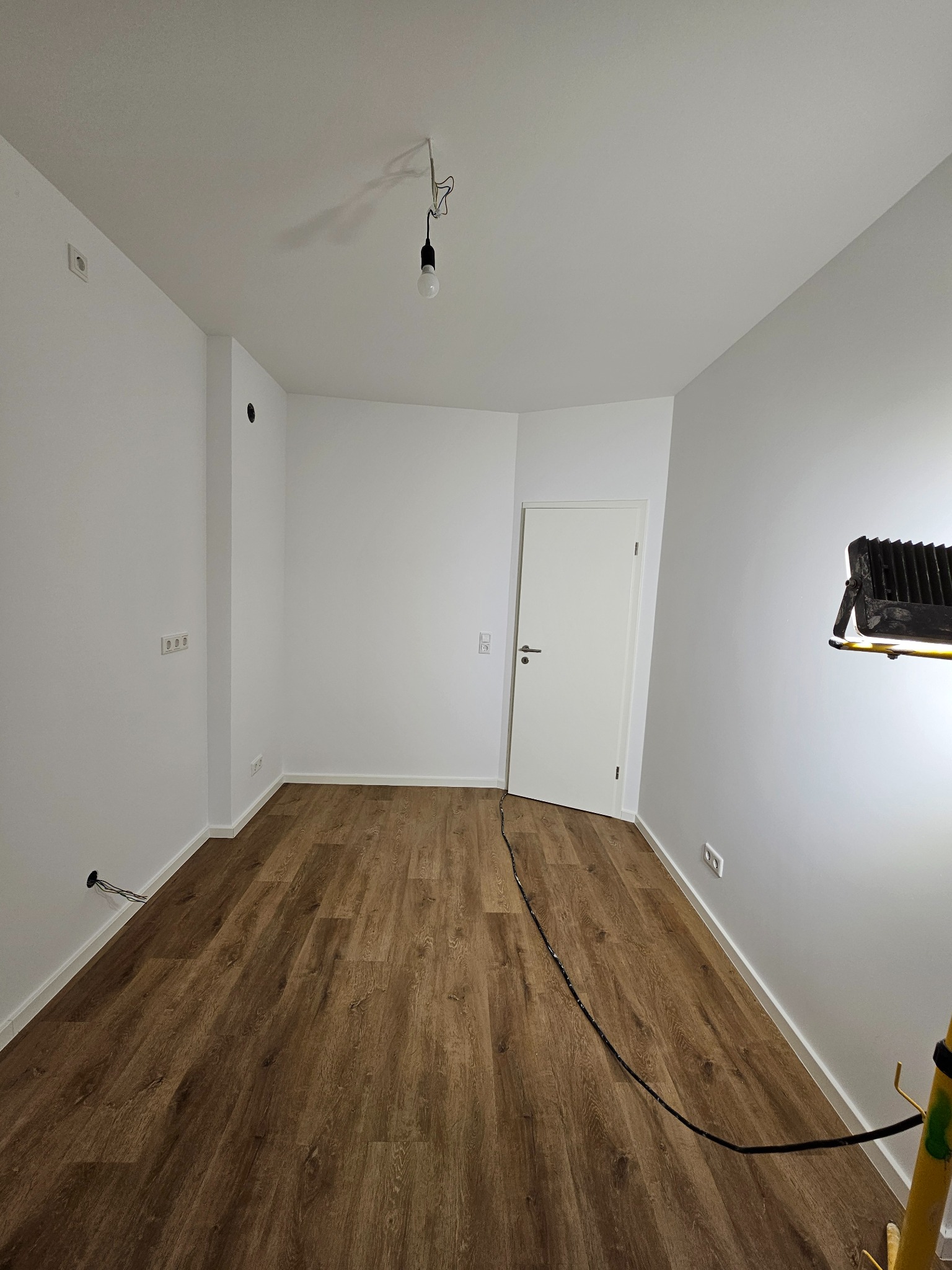 Pokój po remoncie: brązowe panele podłogowe, białe ściany, białe drzwi. Widoczne gniazdka elektryczne i kabel na podłodze. Lampa robocza oświetla ścianę.
