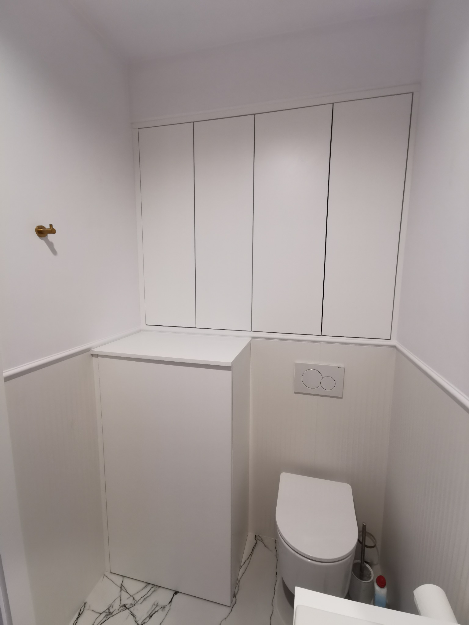 Biała toaleta z zabudową szafek nad stelażem, wykończona w minimalistycznym stylu z marmurową podłogą i złotym haczykiem na ścianie.