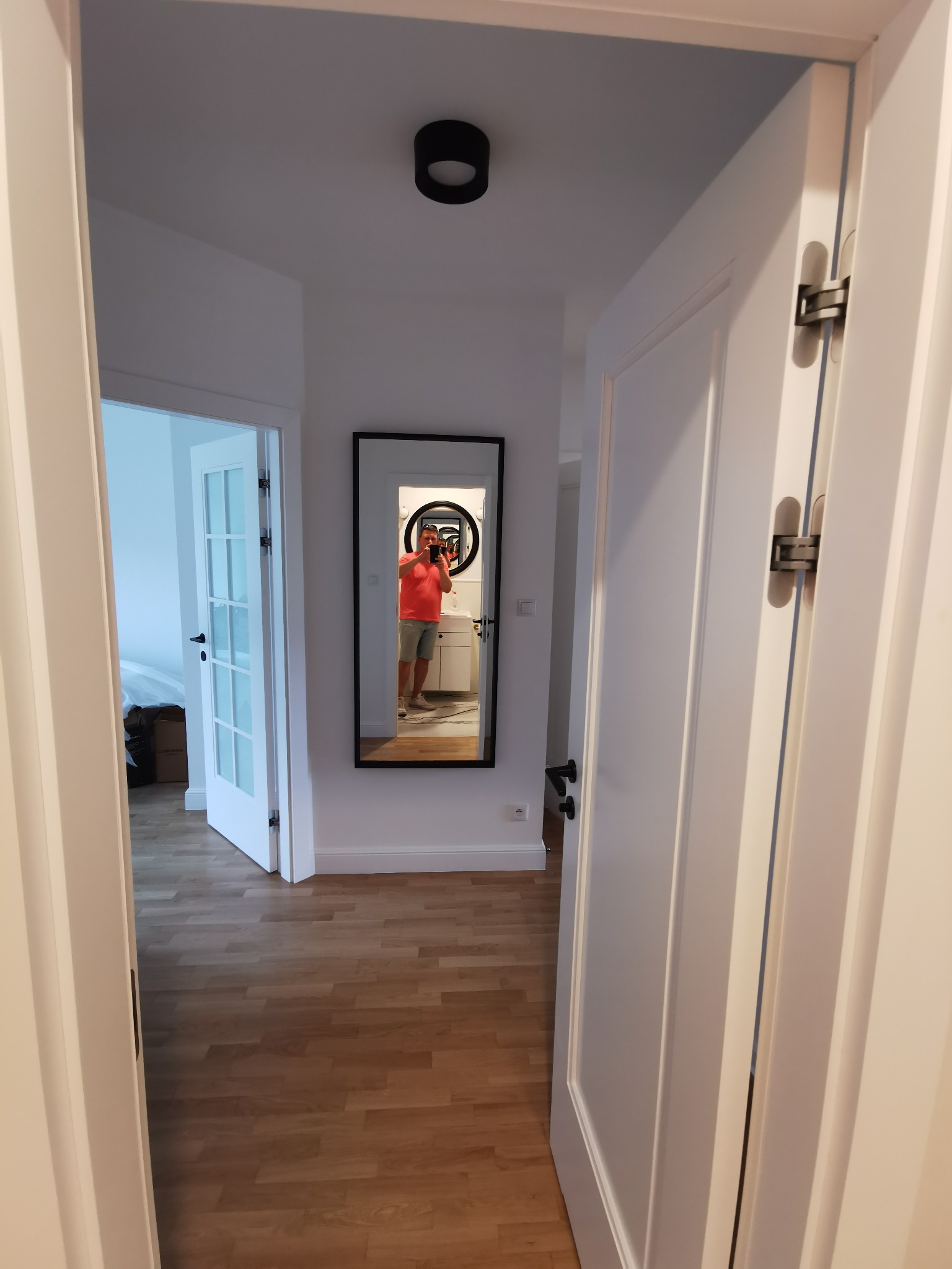 Białe drzwi wewnętrzne z widokiem na korytarz z lustrem, w którym odbija się osoba robiąca zdjęcie. Widoczna podłoga z paneli i drzwi do innego pokoju.