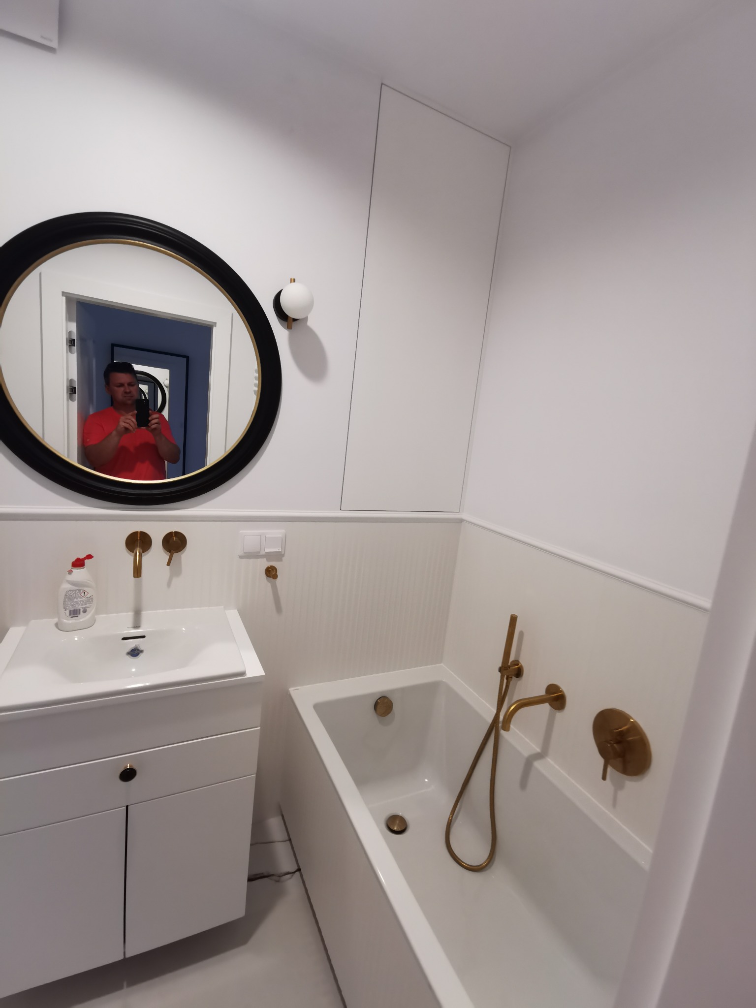 Biała łazienka z wanną i umywalką, złota armatura, okrągłe lustro z czarną ramą. W lustrze odbija się osoba robiąca zdjęcie.