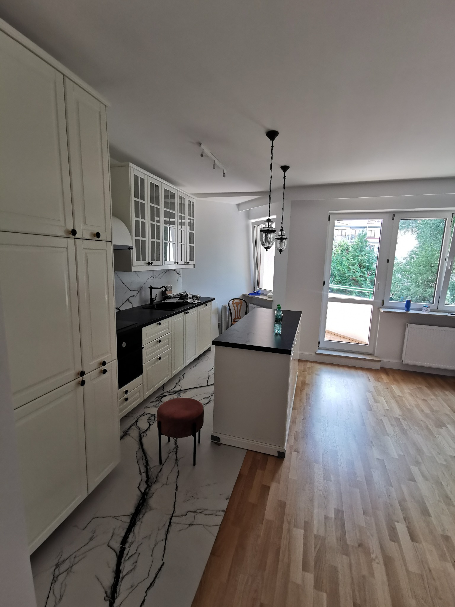 Nowoczesna, jasna kuchnia z wyspą, czarnymi blatami i marmurową podłogą. Widoczne okno z widokiem na zieleń i drewniana podłoga w dalszej części pomieszczenia.