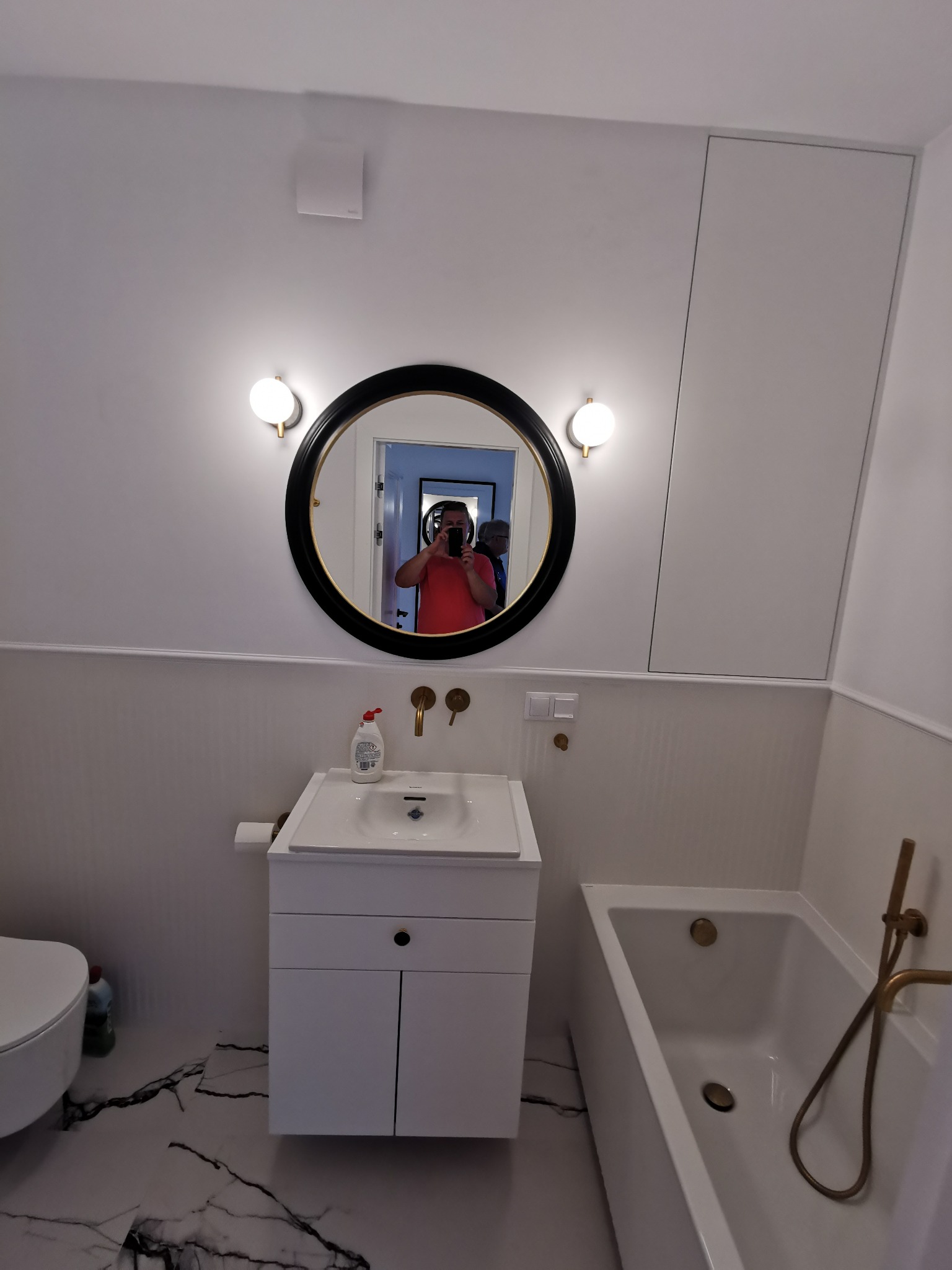 Biała łazienka z okrągłym lustrem, w którym odbija się osoba robiąca zdjęcie. Marmurowe płytki podłogowe i złote akcenty armatury dodają elegancji.