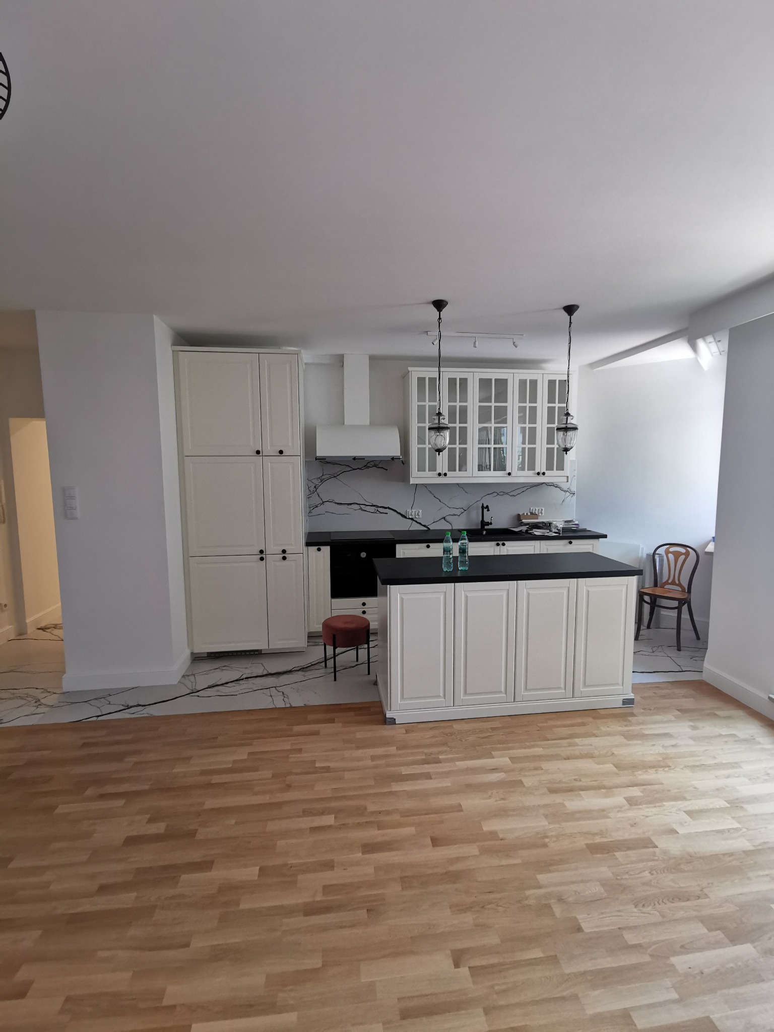 Jasna kuchnia z wyspą i marmurowym akcentem na ścianie, widok na drewnianą podłogę. Nowoczesne wykończenie wnętrza z dbałością o detale.