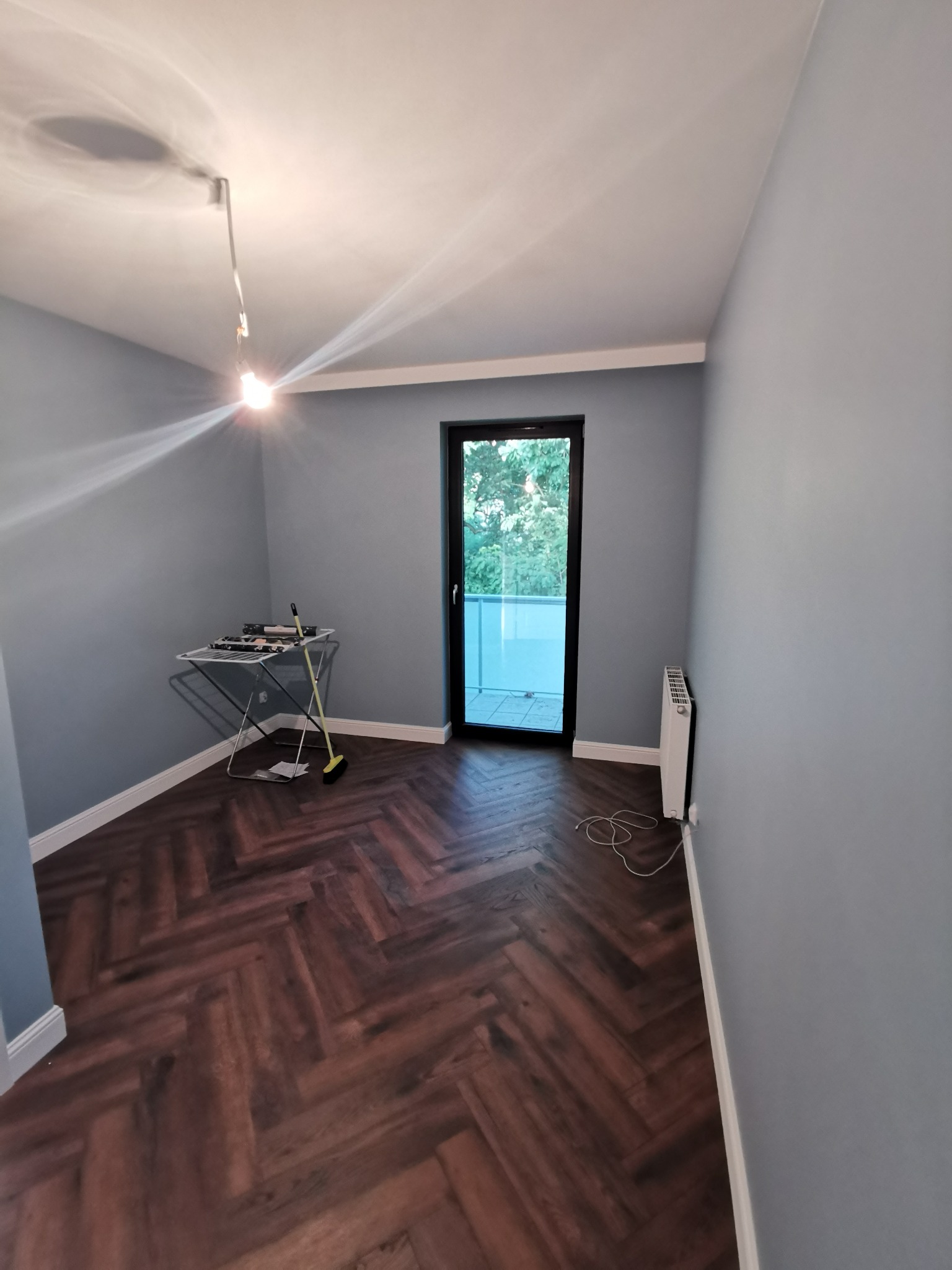 Pokój po remoncie z parkietem w jodełkę, świeżo pomalowanymi szarymi ścianami i wyjściem na balkon. Widoczny grzejnik i suszarka na pranie.