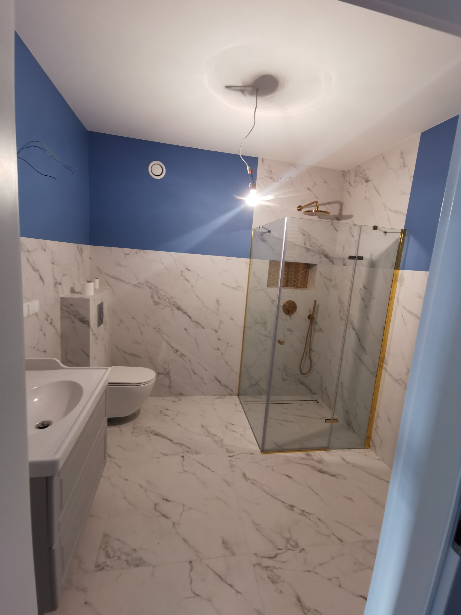 Wykończona łazienka z kabiną prysznicową w złotych ramach, marmurowymi płytkami i niebieskimi akcentami. Widoczna umywalka z szafką i toaleta.
