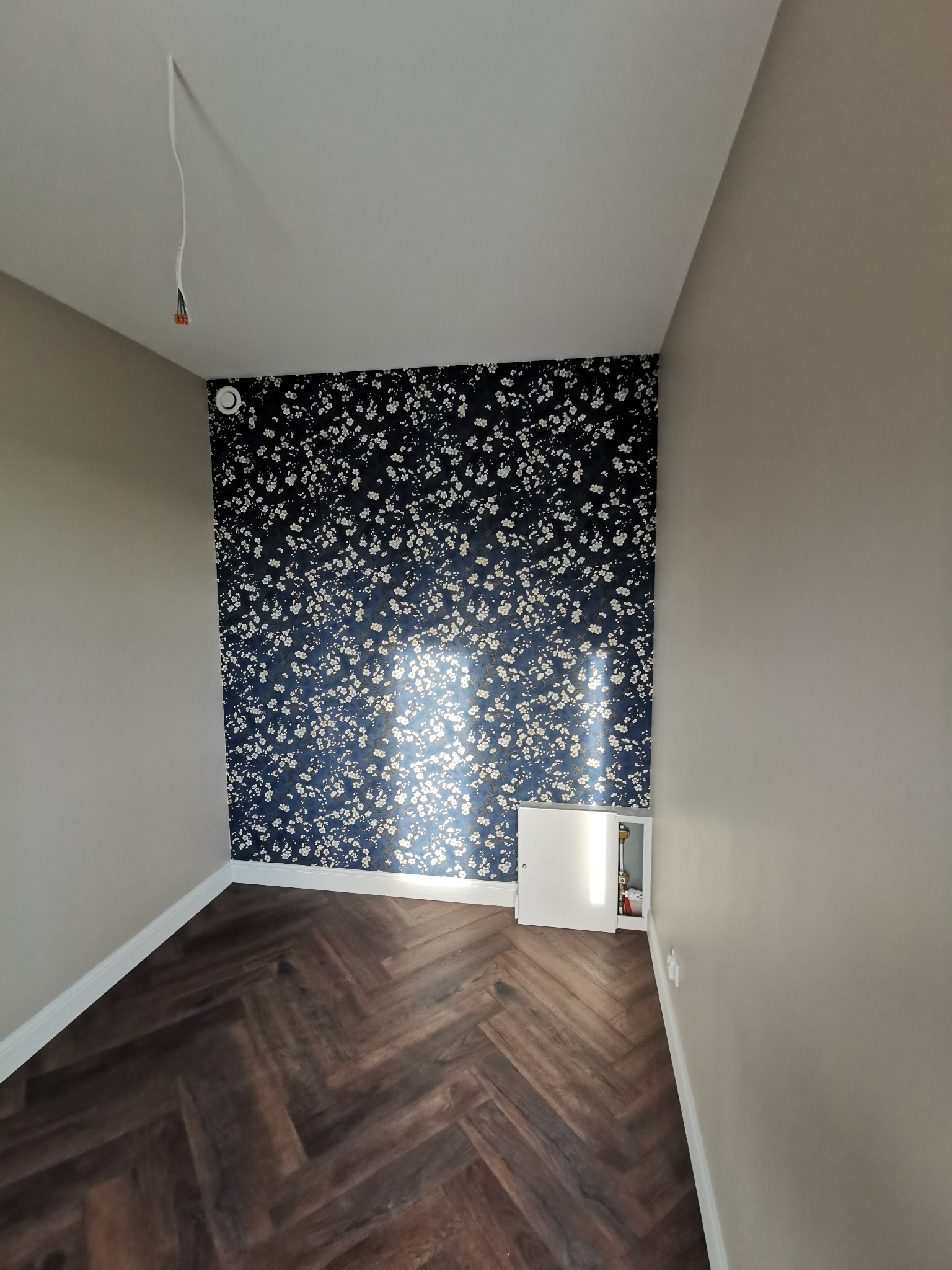 Pokój po remoncie: parkiet w jodełkę, ściana z tapetą w kwiaty, gładkie ściany w neutralnym odcieniu, widoczne kable wystające z sufitu, biała klapka rewizyjna.