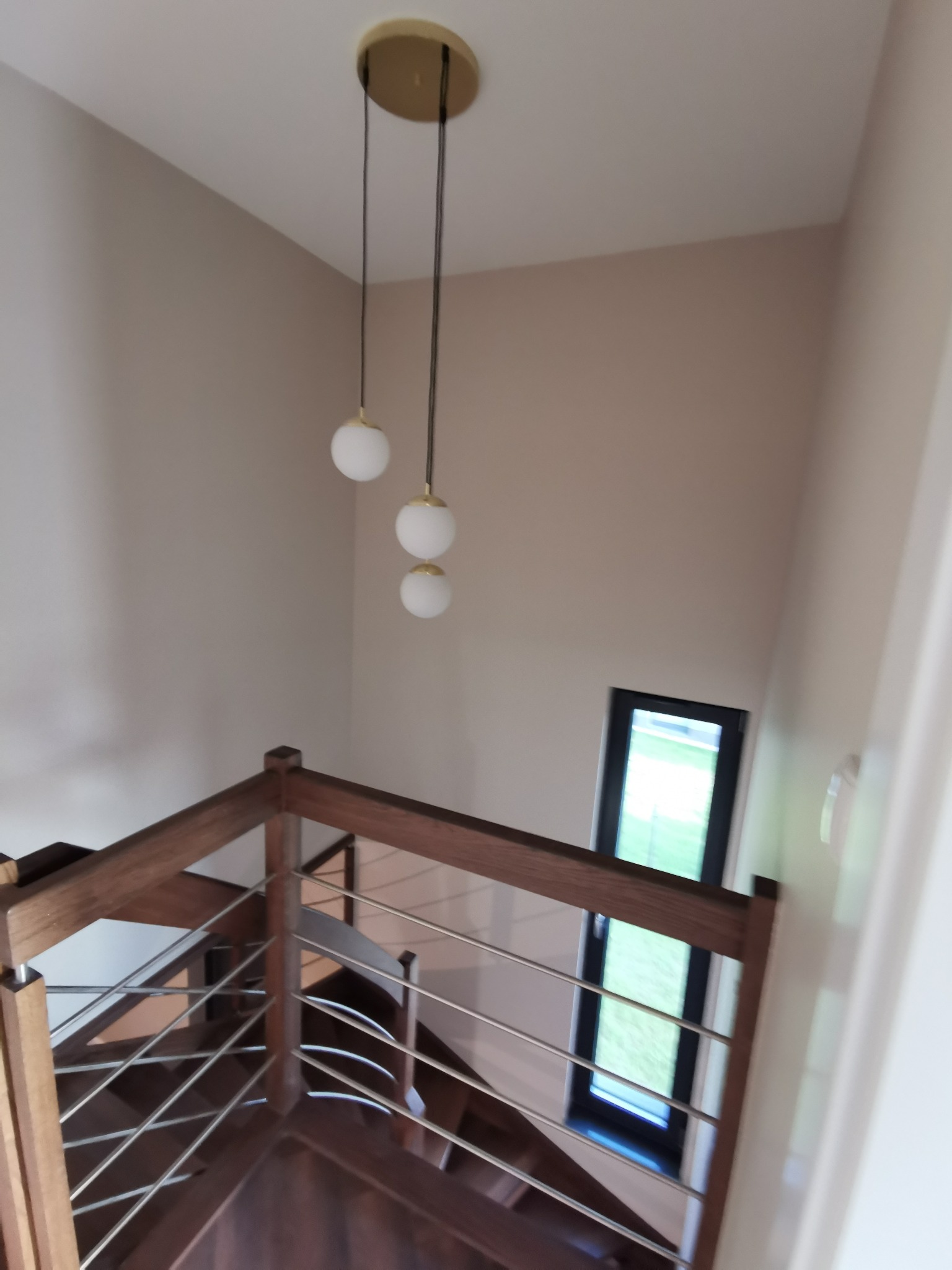 Nowoczesne schody z drewnianymi stopniami i metalową balustradą, oświetlone wiszącą lampą z trzema kulistymi kloszami, widok z góry na tle okna.
