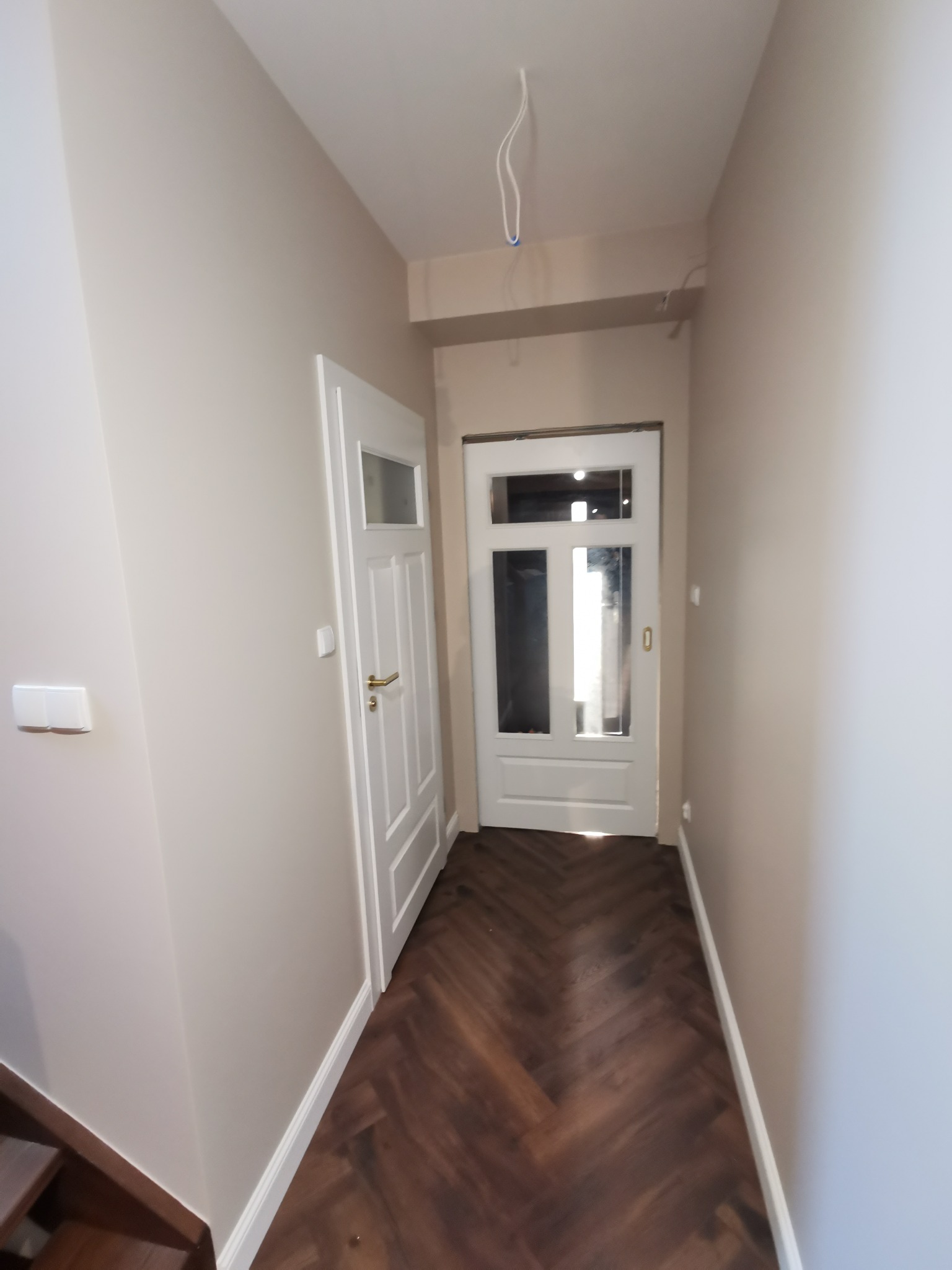 Korytarz po remoncie: parkiet w jodełkę, świeżo pomalowane ściany w odcieniach beżu, białe drzwi z przeszkleniami i widoczne kable oświetleniowe.