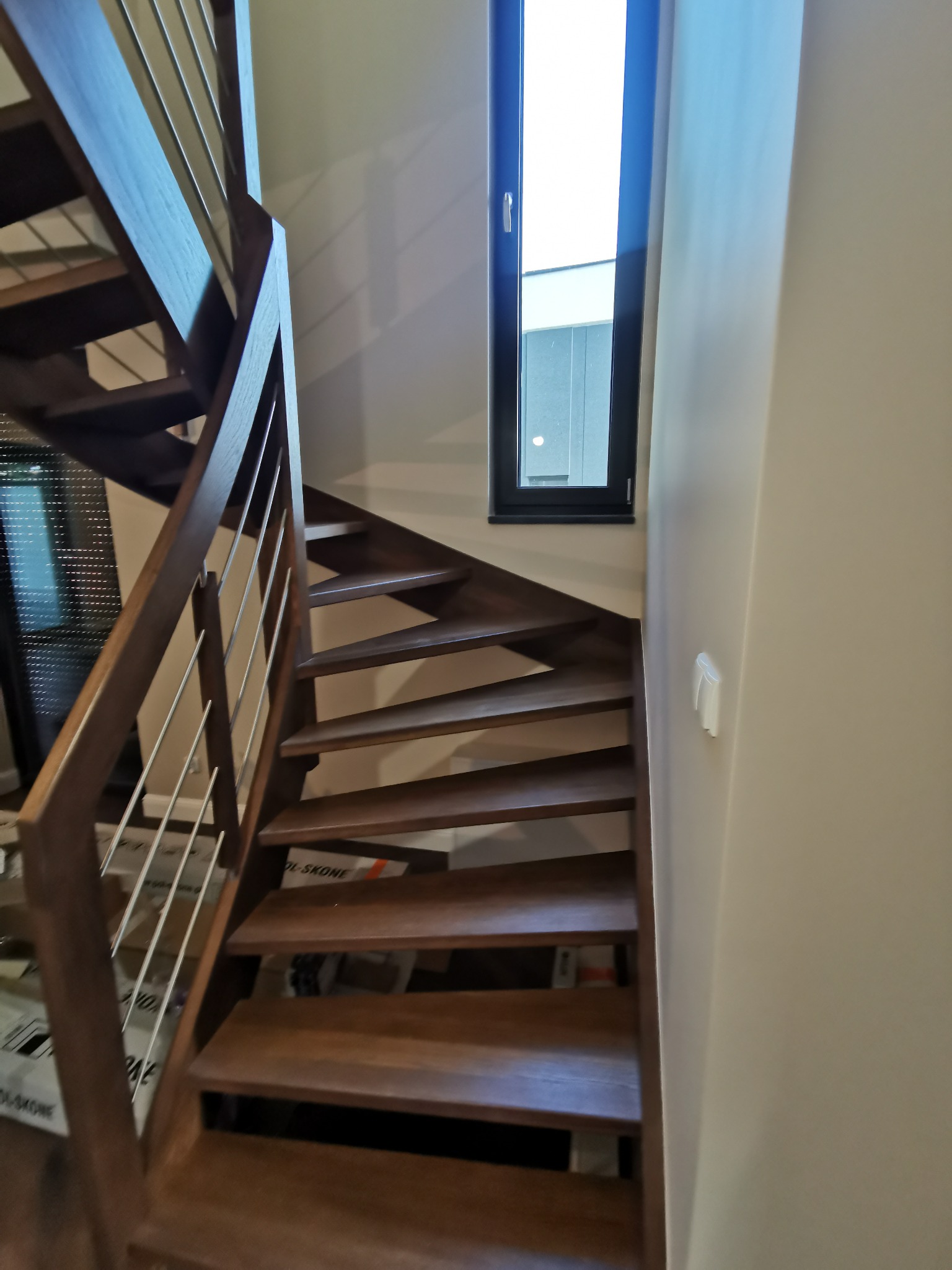 Nowoczesne, drewniane schody z metalową balustradą, oświetlone naturalnym światłem z okna. Minimalistyczny design, ciepłe odcienie drewna, jasne ściany.