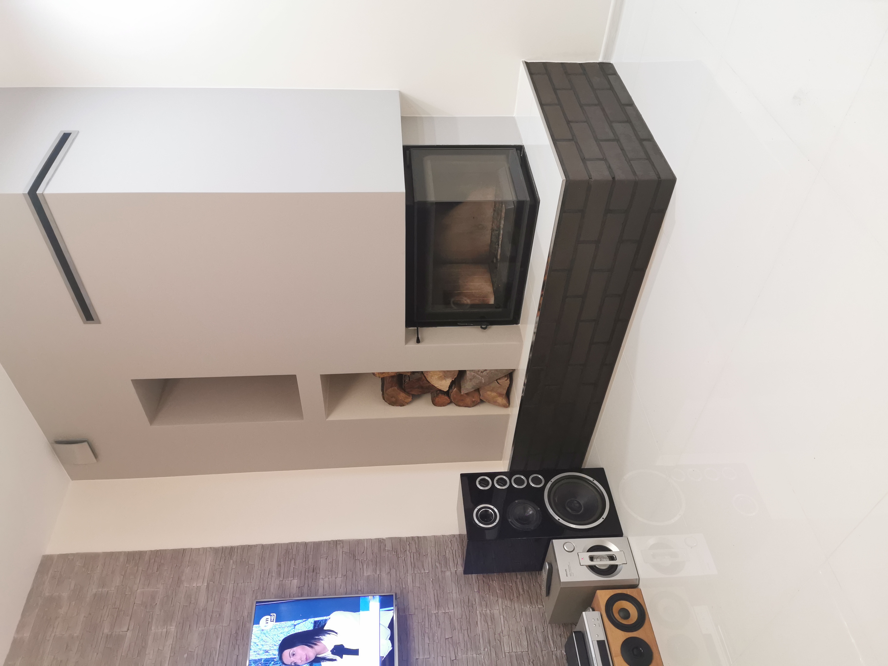 Nowoczesny kominek z czarną obudową i wkładem na drewno, obok ściana wykończona w czarno-szarą cegłę, w tle fragment ściany z dekoracyjnym kamieniem i telewizorem.