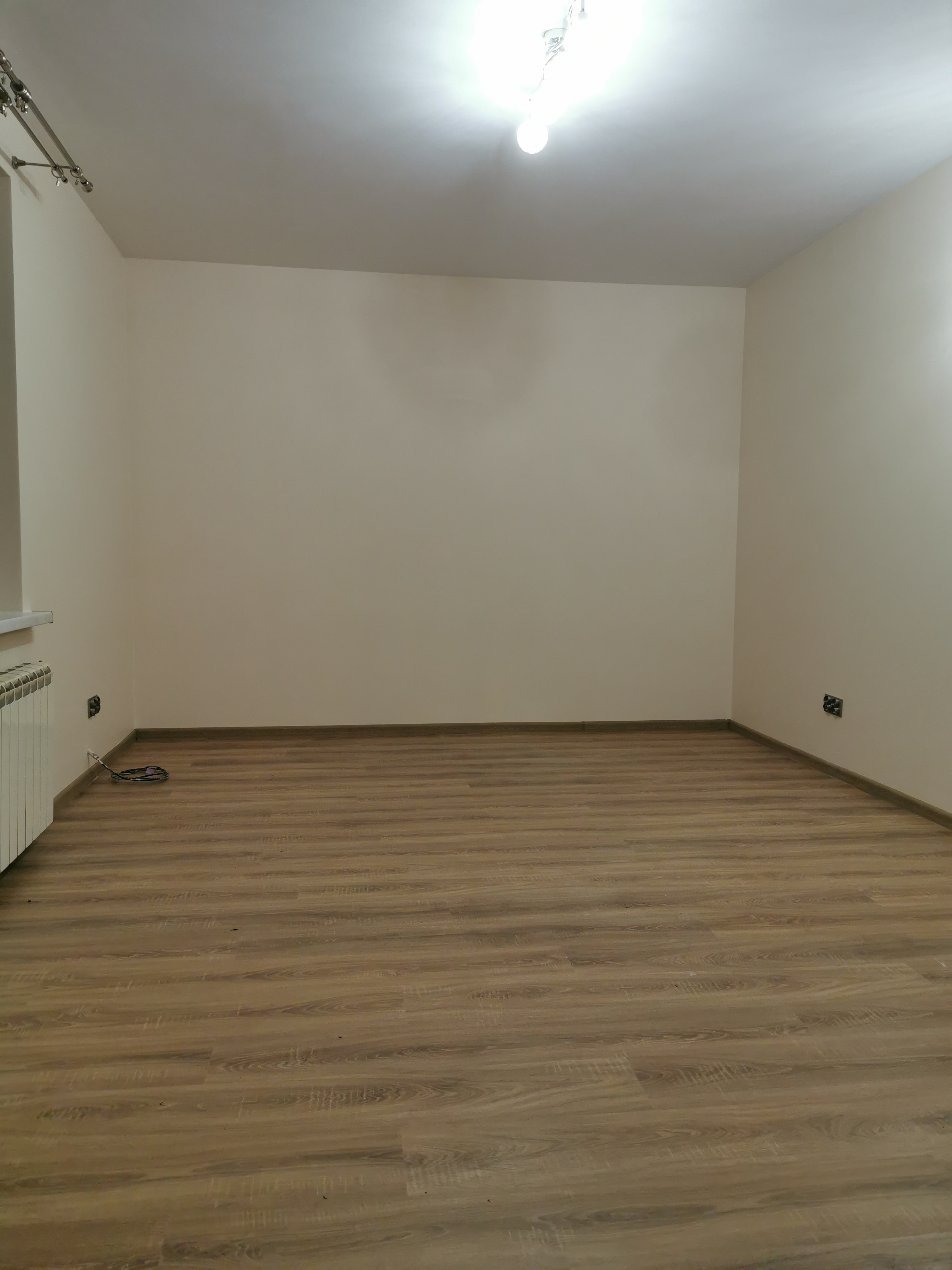 Pusty pokój po remoncie z panelami podłogowymi w odcieniach brązu, świeżo pomalowanymi ścianami w kolorze beżowym, grzejnikiem i wystającą instalacją elektryczną.