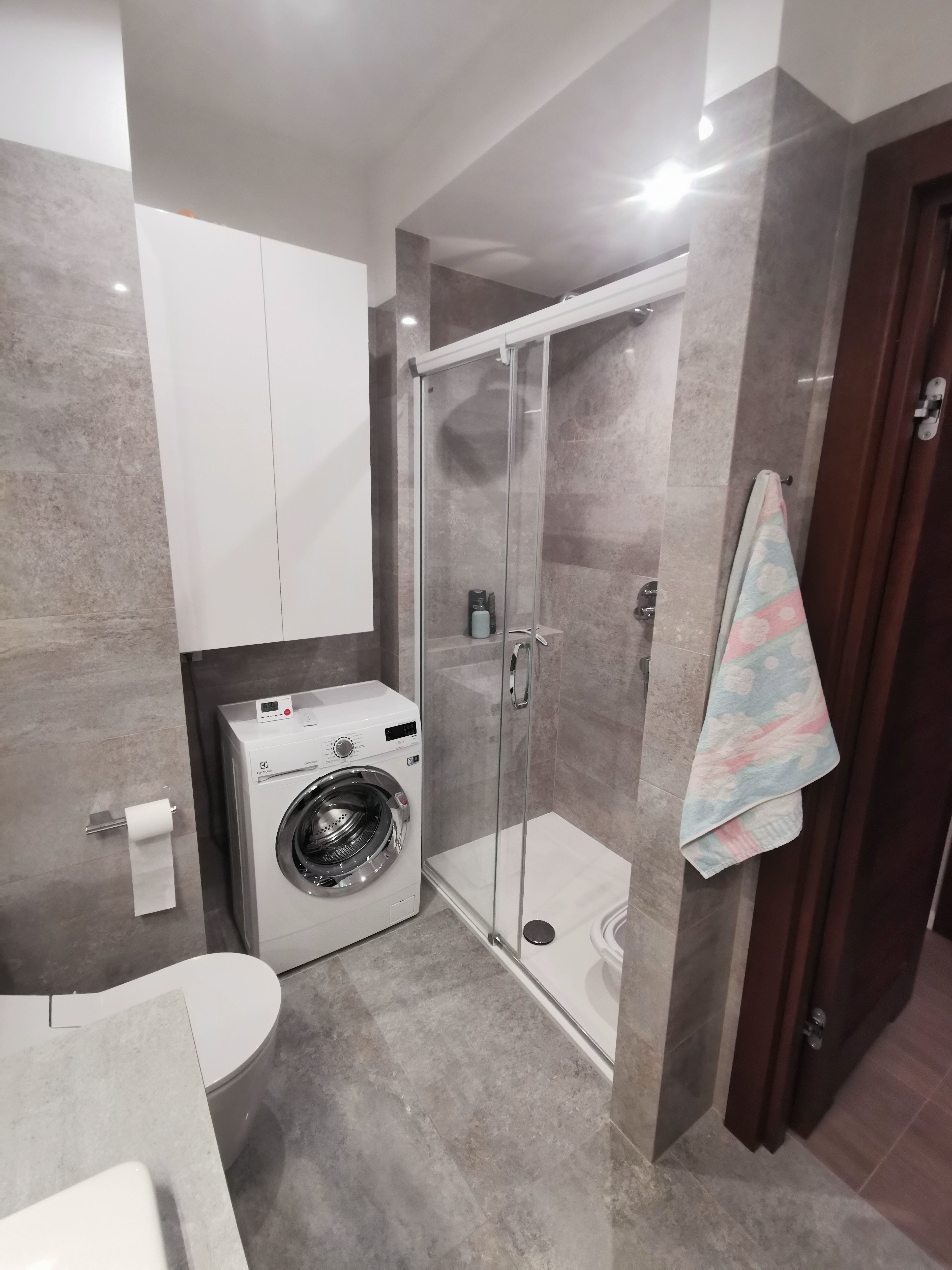 Nowoczesna łazienka z szarą płytką imitującą kamień, białą szafką wiszącą, pralką Electrolux, kabiną prysznicową ze szklanymi drzwiami przesuwnymi i grzejnikiem drabinkowym.