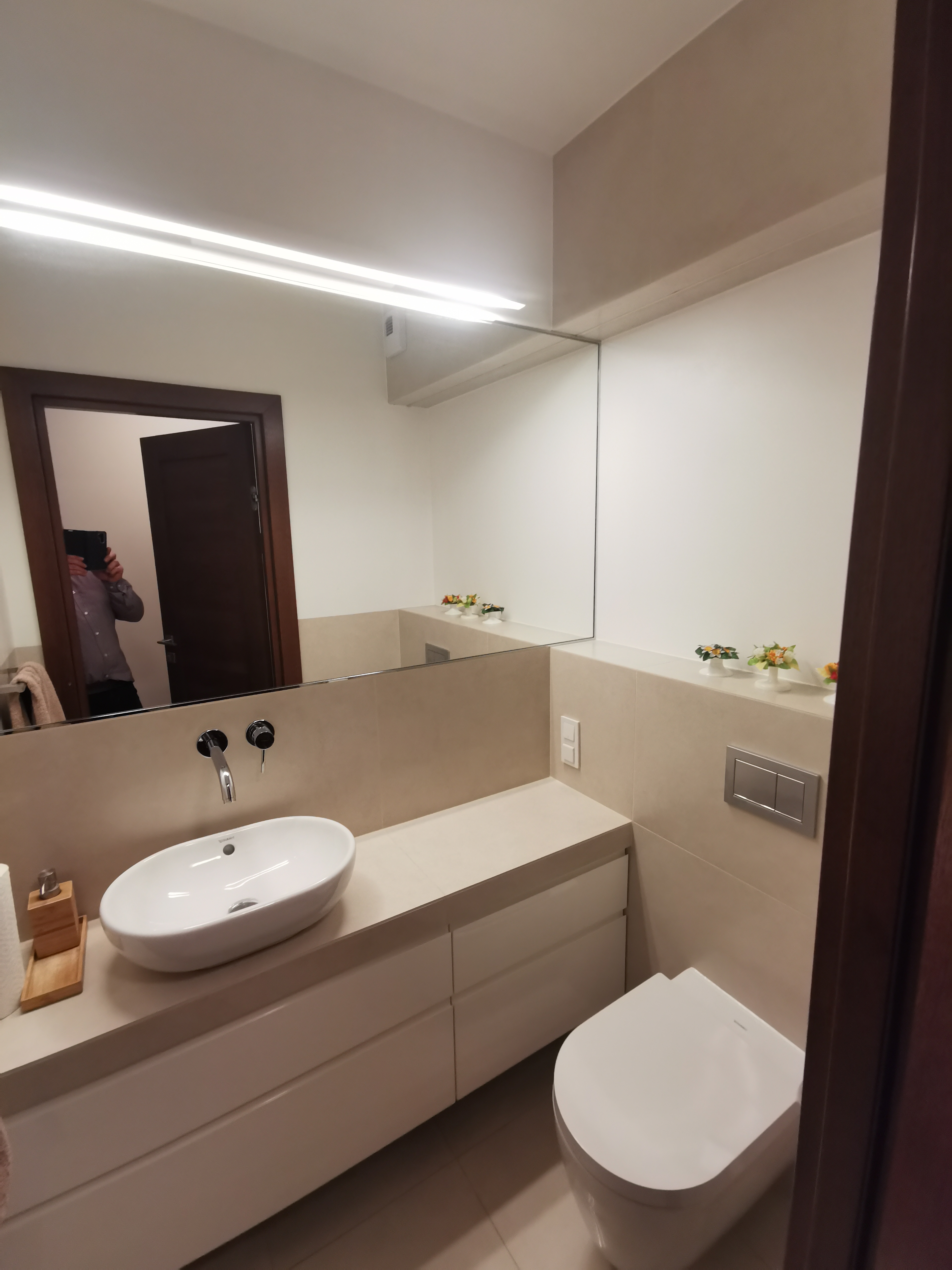Nowoczesna łazienka z owalną umywalką nablatową, baterią ścienną, podświetlanym lustrem, wiszącą toaletą i beżowymi płytkami ściennymi. W lustrze odbija się osoba robiąca zdjęcie.