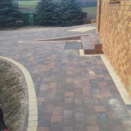 KABUD Karol Sikora - Ułożona z kostki brukowej ścieżka prowadząca do domu z widocznymi schodami i rampą, obok trawnika z psem rasy doberman w lewym dolnym rogu.